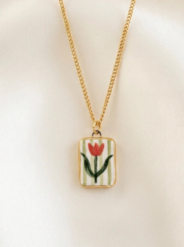 Gold necklace with a pendant featuring a red tulip design and green stripes on a beige background.