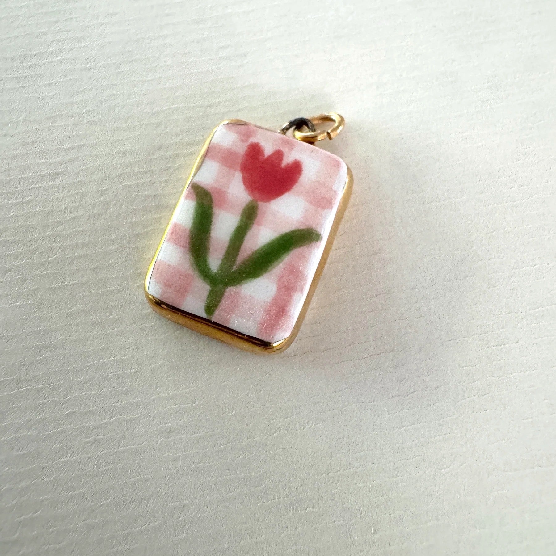 Small rectangular pendant with a red flower design on a pink checkered background on light grey background.