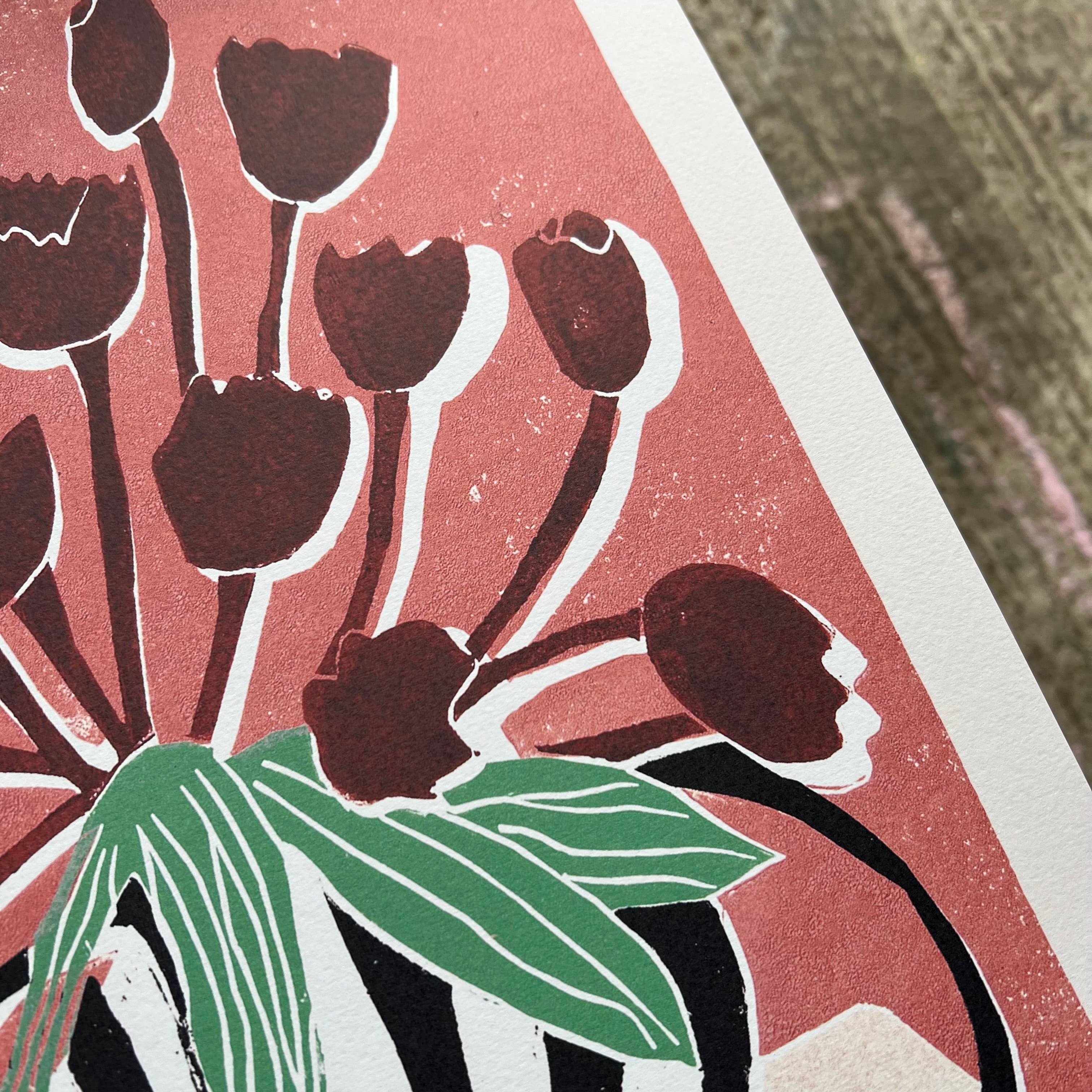 A close-up of a print featuring dark red tulip buds with green leaves on a pink background, bordered by a white sheet of paper.