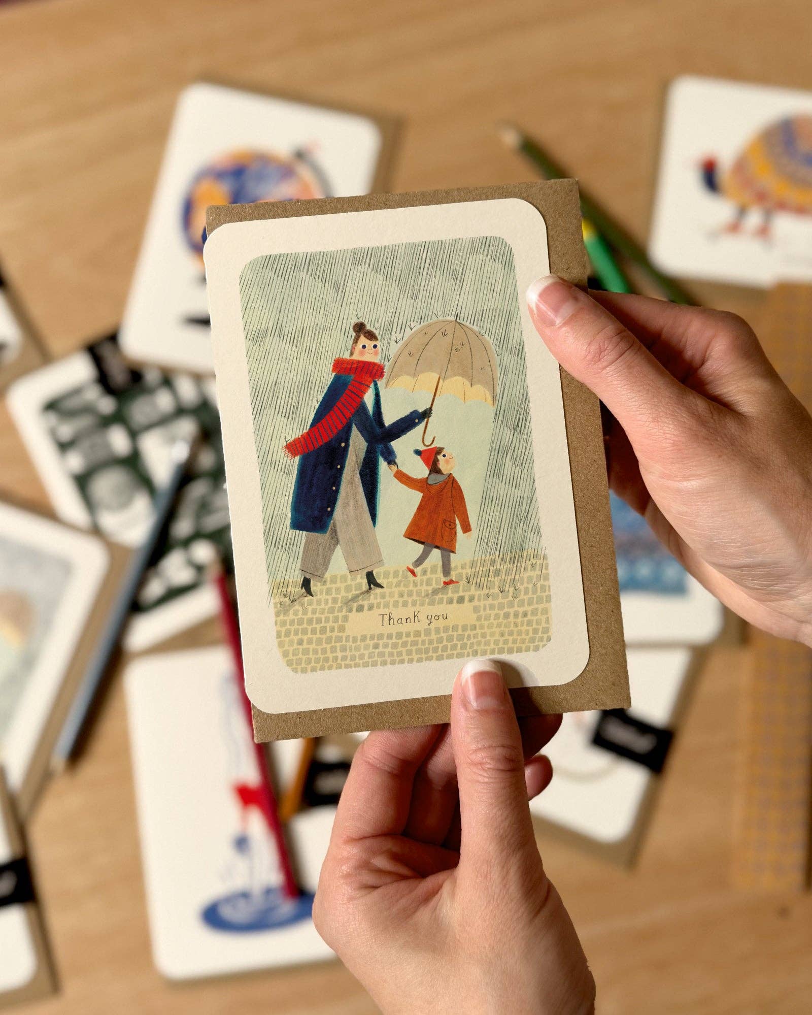 Hand holding a card with an illustration of a couple under an umbrella, surrounded by other cards on a wooden surface.