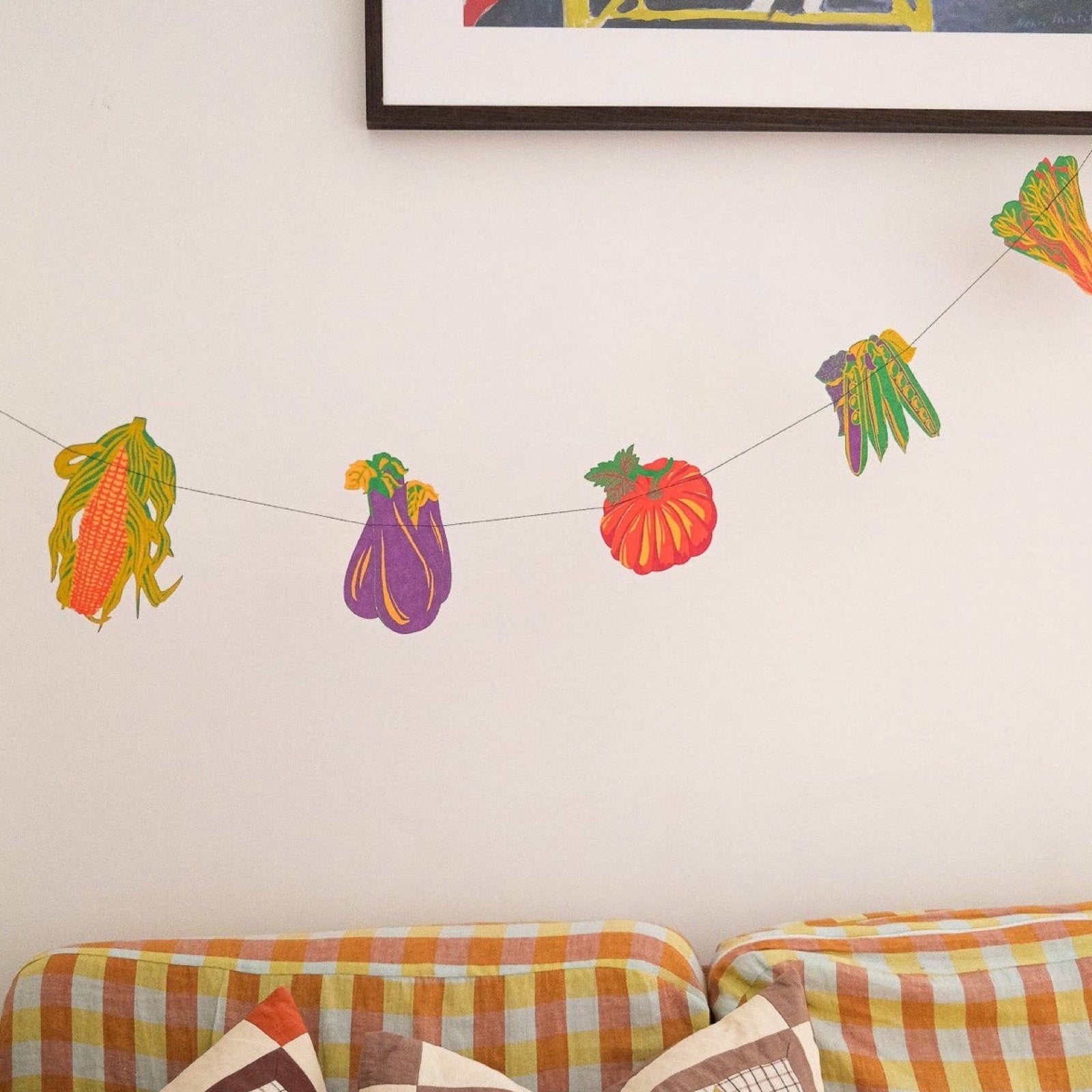 A vegetable garland with twelve screen-printed vegetables including carrots, beetroot, sweetcorn, asparagus and eggplant hanging on a white wall above an orange checked sofa.