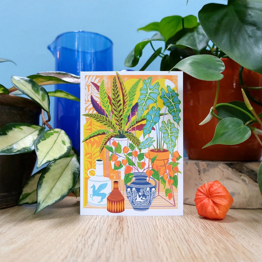 Colorful card with plant illustrations surrounded by potted plants on a wooden surface.