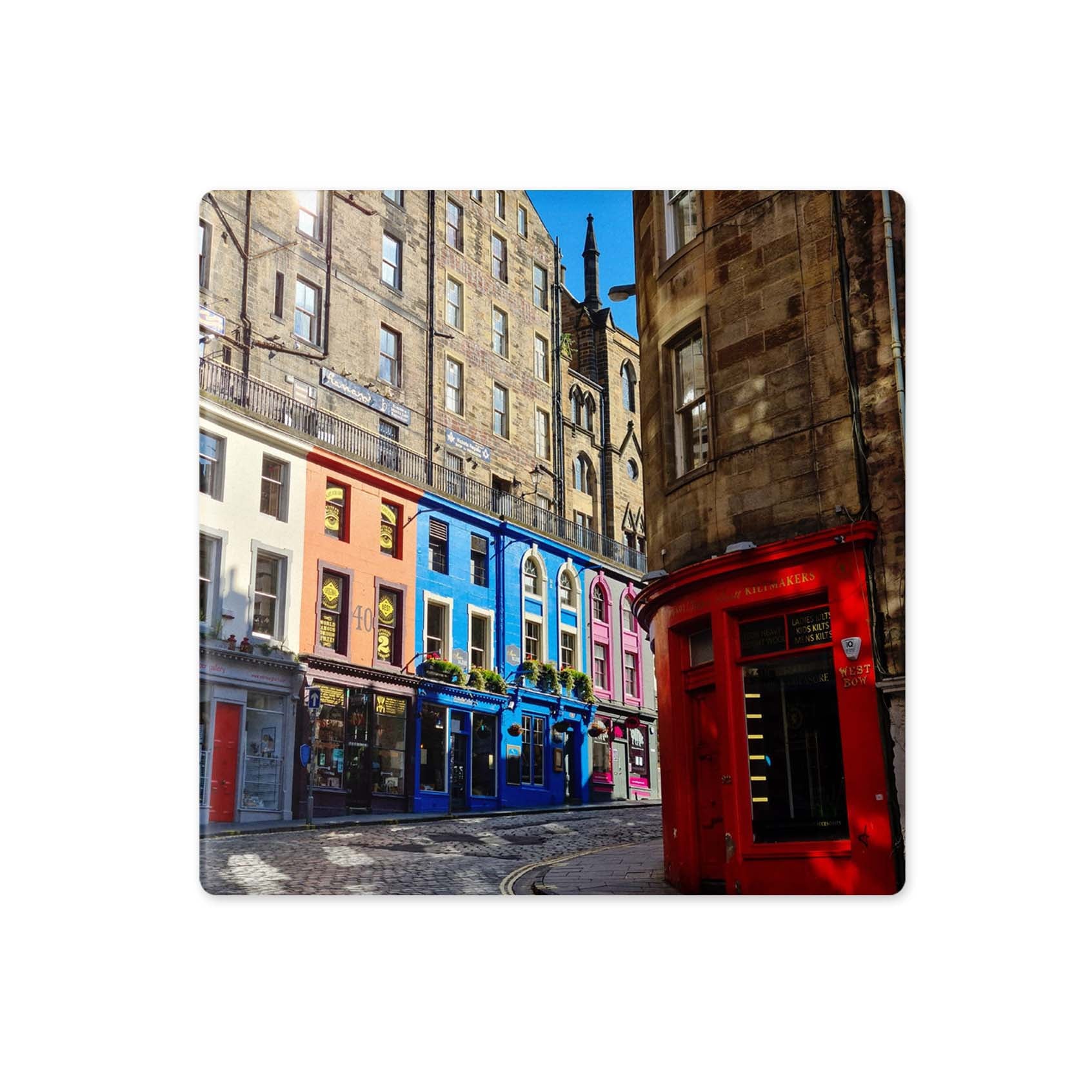 A square coaster featuring a colorful photograph of a street in Edinburgh, Scotland, with vibrant blue and pink buildings, a red phone booth, and historic stone towers under a blue sky.