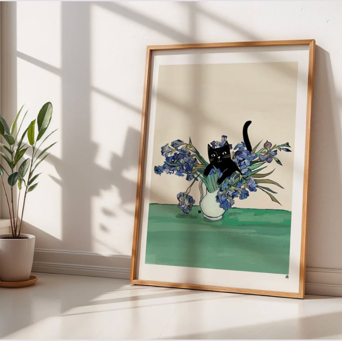Framed artwork of a black cat among flowers on a green surface, sat on the floor with a plant in the background.