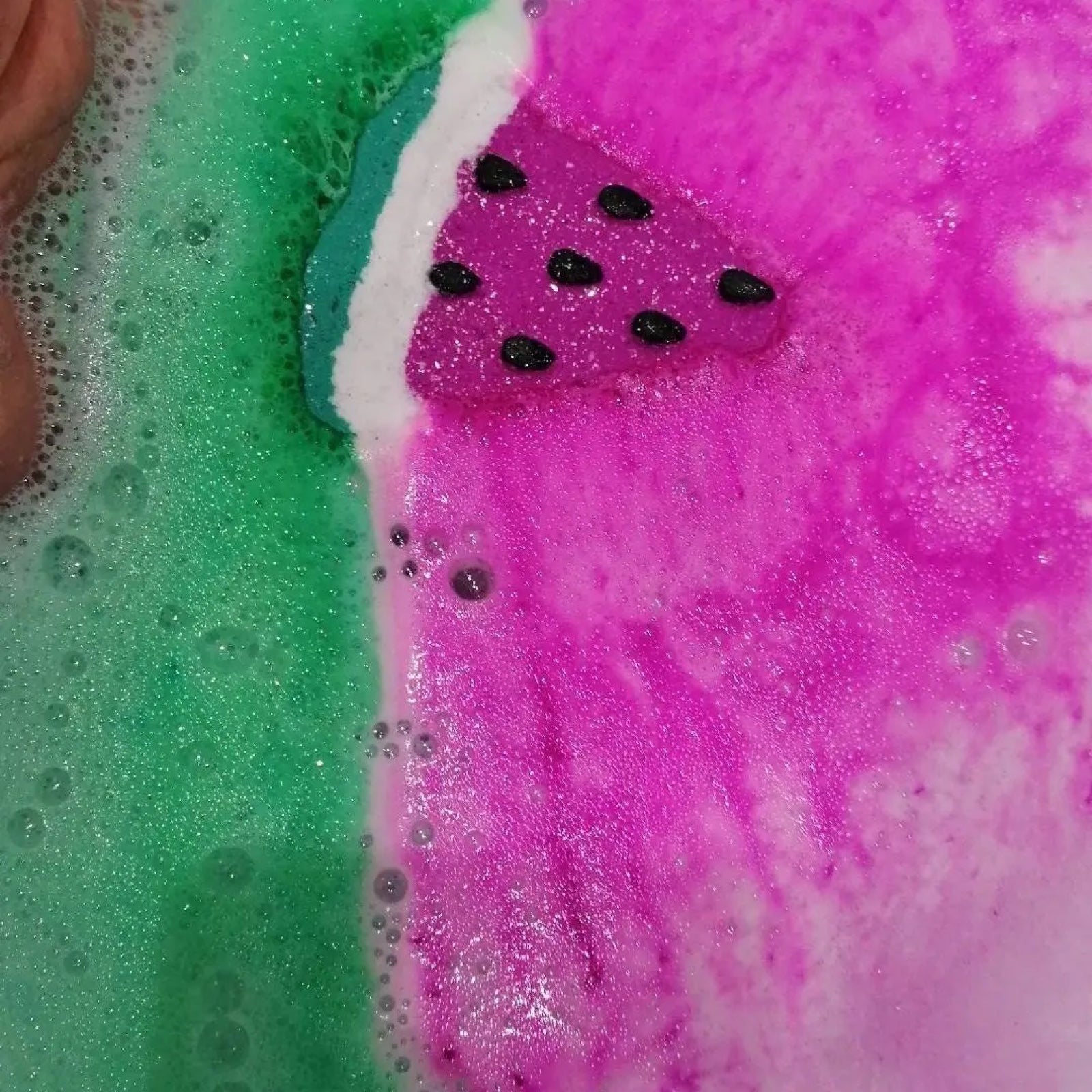 A pink, green and white watermelon slice-shaped bath bomb dissolving in the bath water and turning it pink and green.