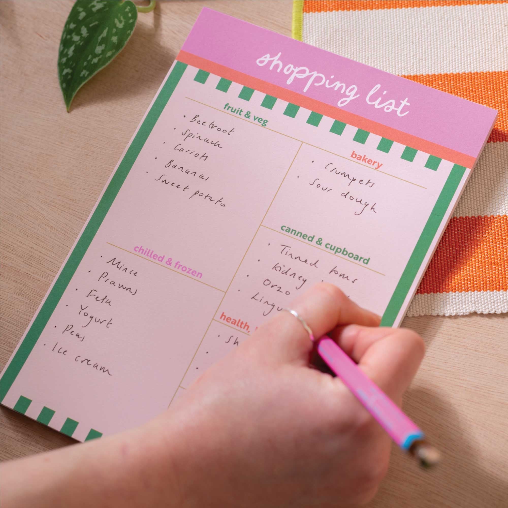 Hand writing on a pink shopping list with a pen, placed on a wooden surface.