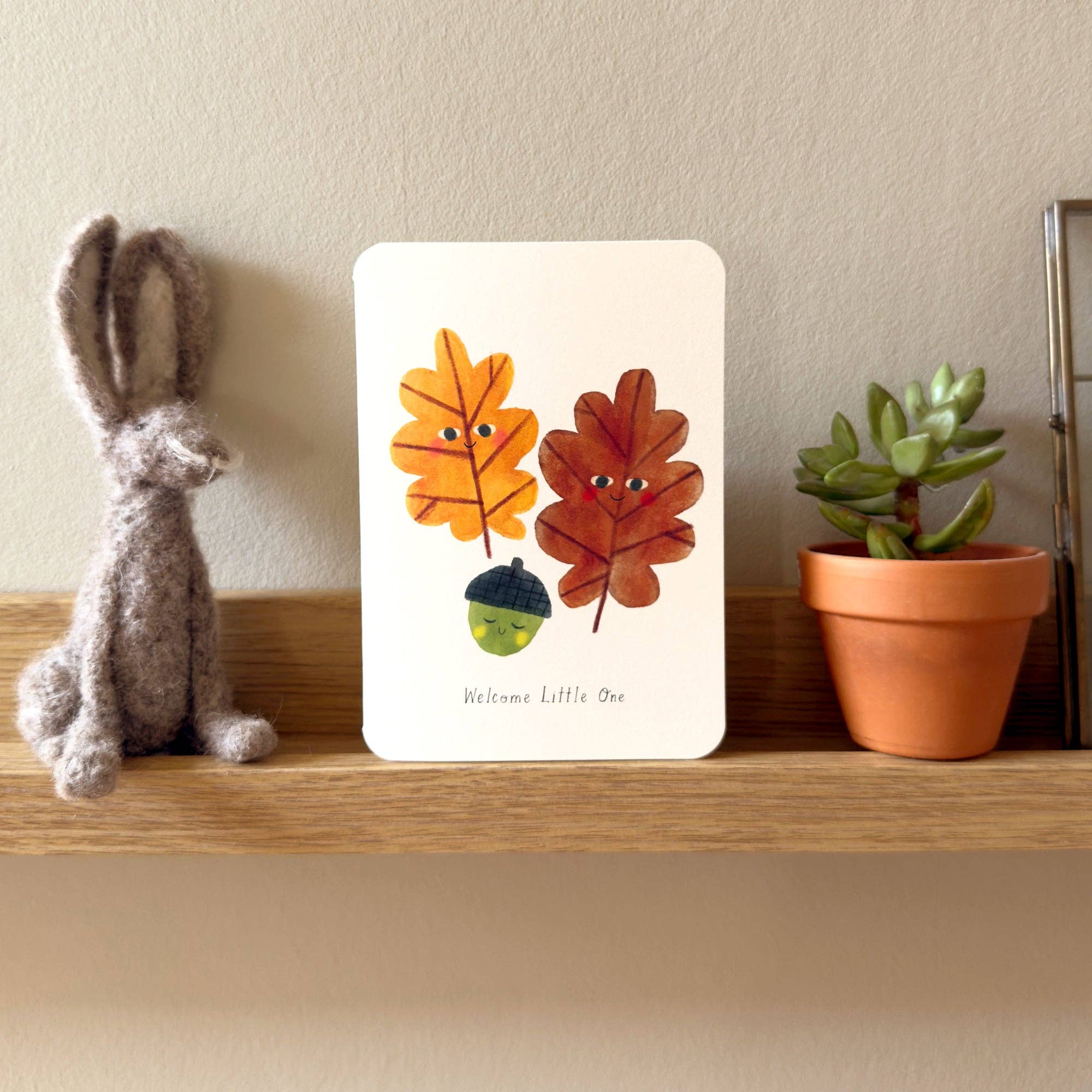 Decorative shelf with a plush rabbit, card with leaf design, and potted plant against a beige wall.
