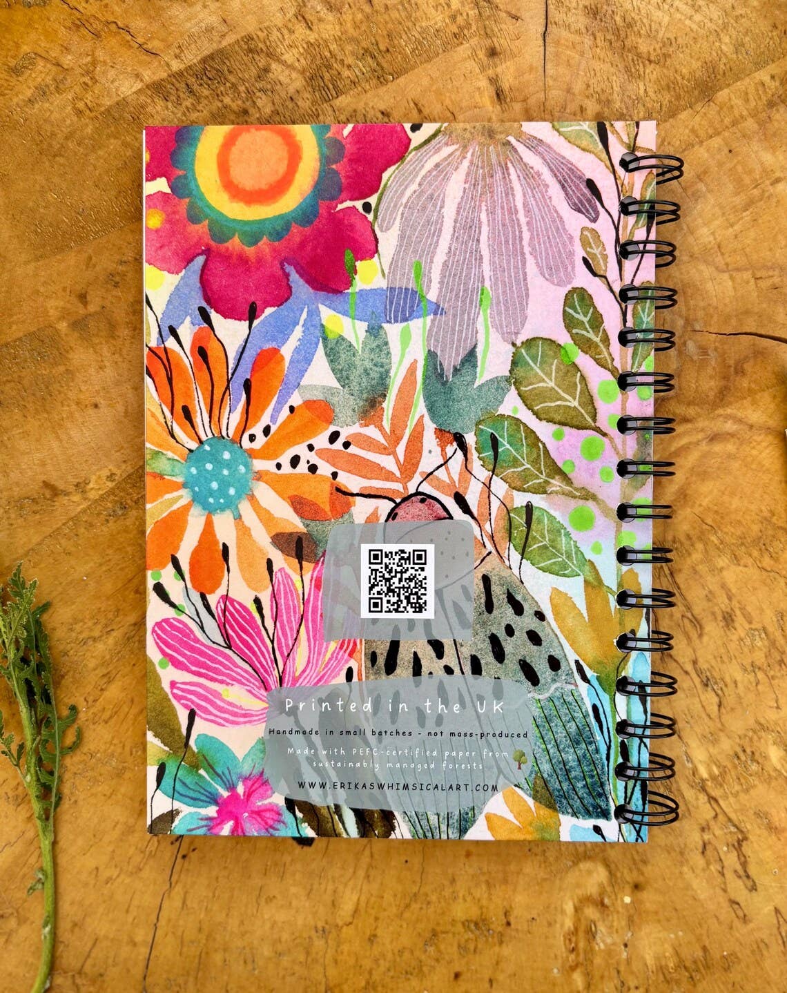 A spiral-bound notebook with a colourful floral and botanical cover design, photographed from above on a rustic wooden surface. The cover features a variety of vibrant flowers, leaves, and abstract patterns in pink, orange, yellow, green, blue, and purple, with a black spiral binding on the right side.