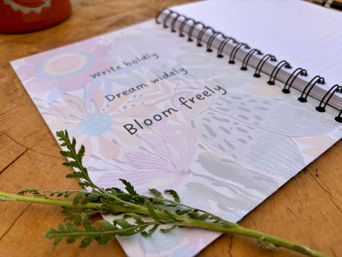 A spiral-bound notebook with a floral cover design reading 'Write boldly. Dream widely. Bloom freely'.