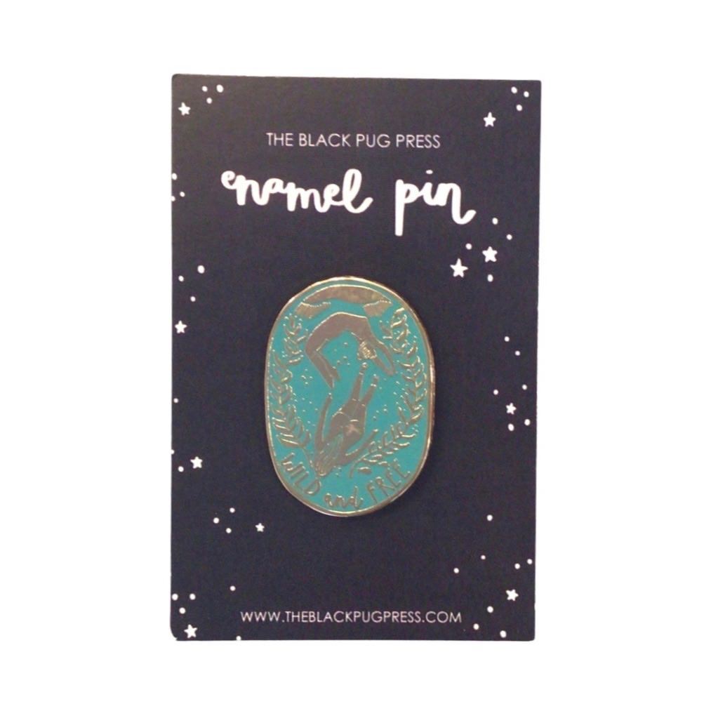 An oval-shaped turquoise enamel pin badge featuring two female wild swimmers and the text 'Wild and Free'.