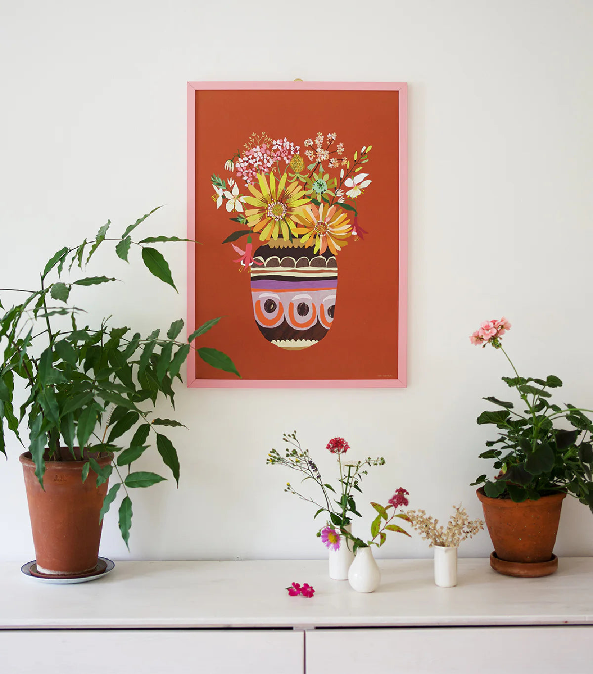 Framed floral artwork on a wall with potted plants below.