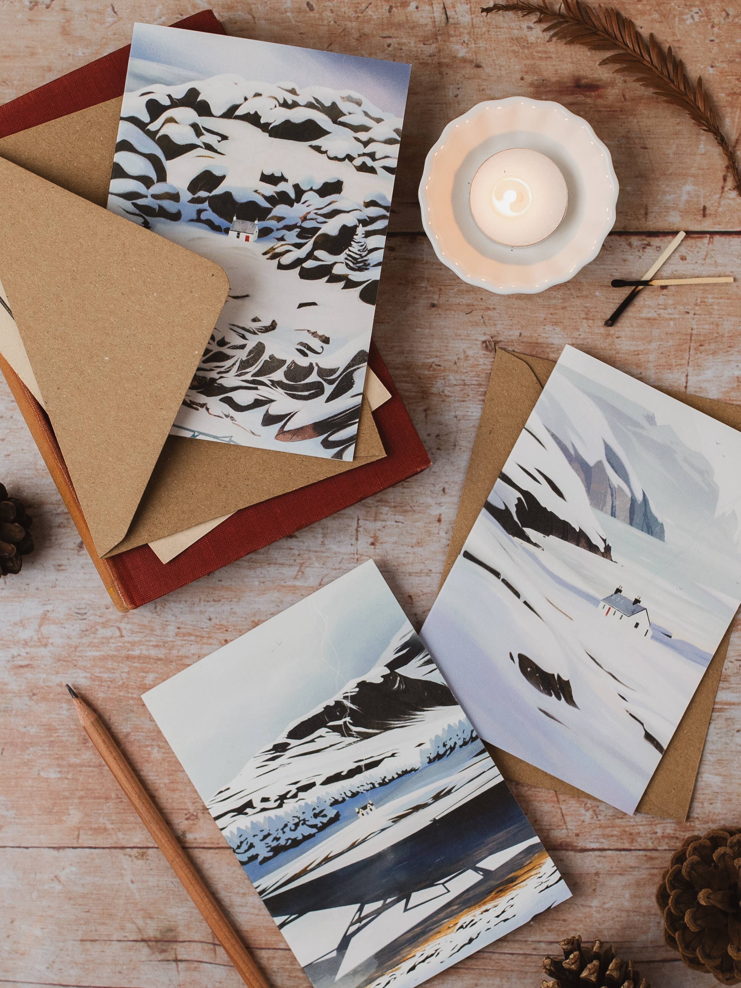 Three art cards featuring winter mountain landscapes in a minimalist black, white, and blue style, arranged on a rustic wooden table with kraft paper envelopes, a lit candle, a pencil, and pinecones.