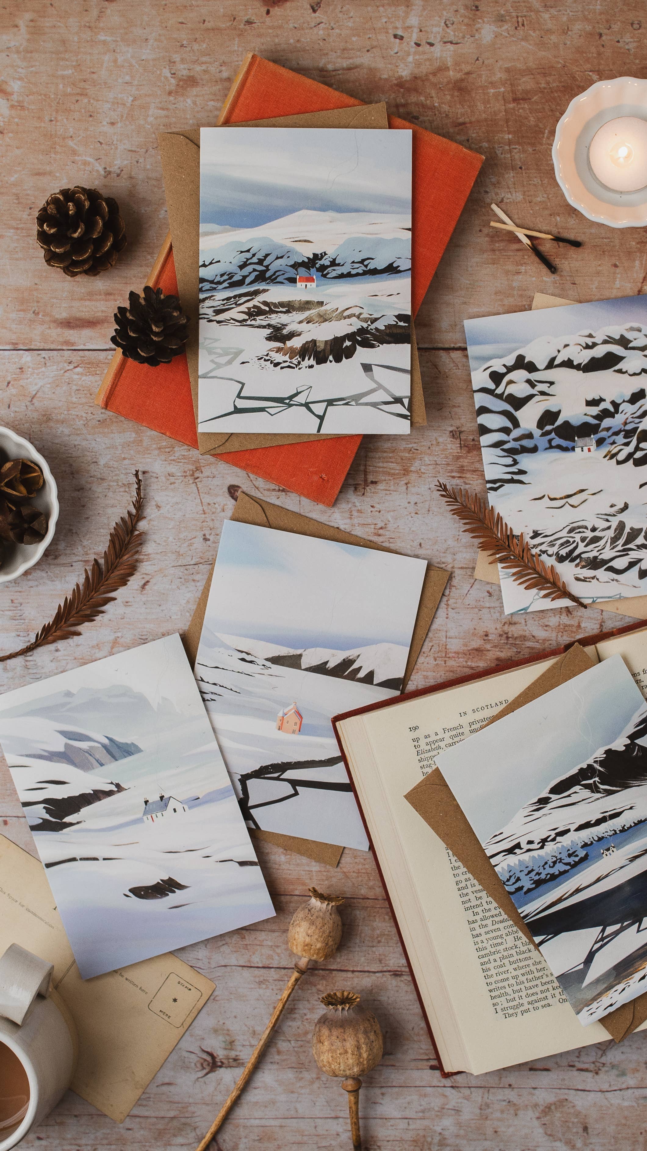 Several cards depicting snowy mountain landscapes, arranged on a rustic wooden table with an open book, pinecones, dried leaves, a candle, and envelopes.