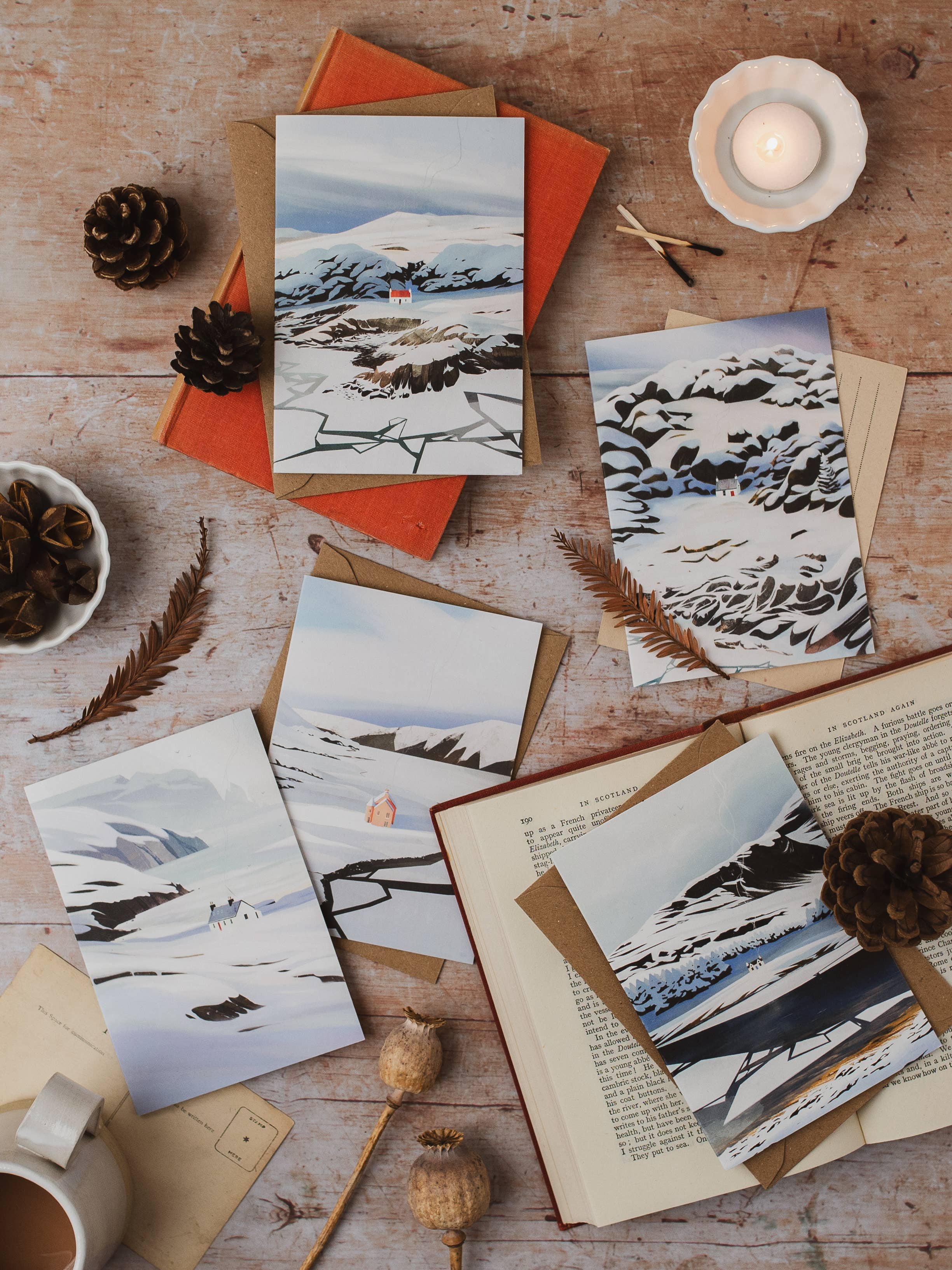 A styled flat-lay of several cards featuring snowy mountain and rocky landscape scenes, arranged on a rustic wooden surface with an open book, pinecones, dried leaves, a candle, and a mug.