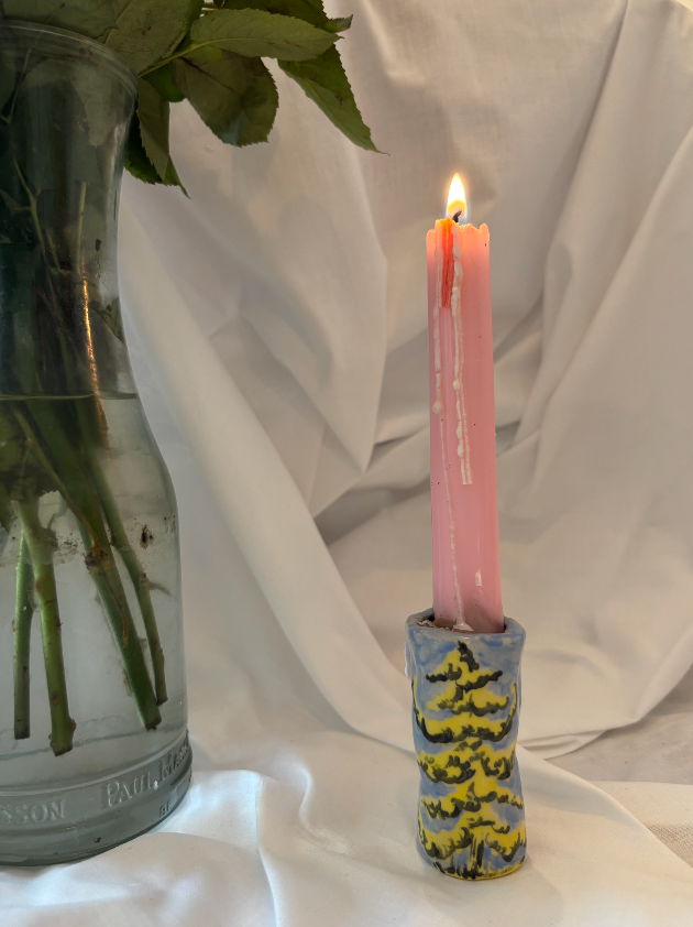 Candle in a holder with a winter tree design, next to a vase of flowers on a white fabric background.