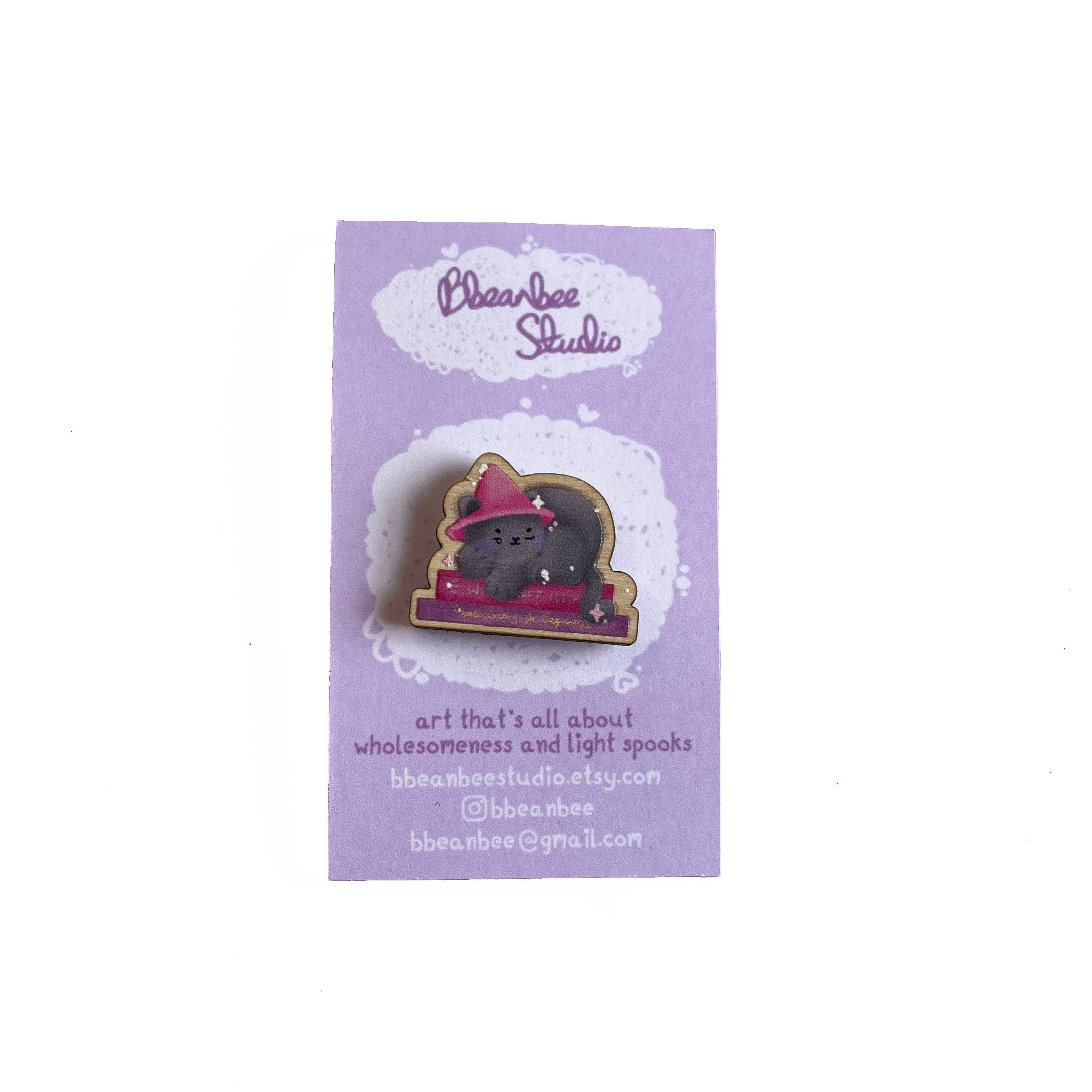 Wooden pin badge of a grey cat wearing a pink witch's hat, sleeping on a stack of books. Book titles are 'Witchcraft 101' and 'Spellcasting for Beginners'. This badge is printed onto 3mm thick responsibly sourced European birch plywood.