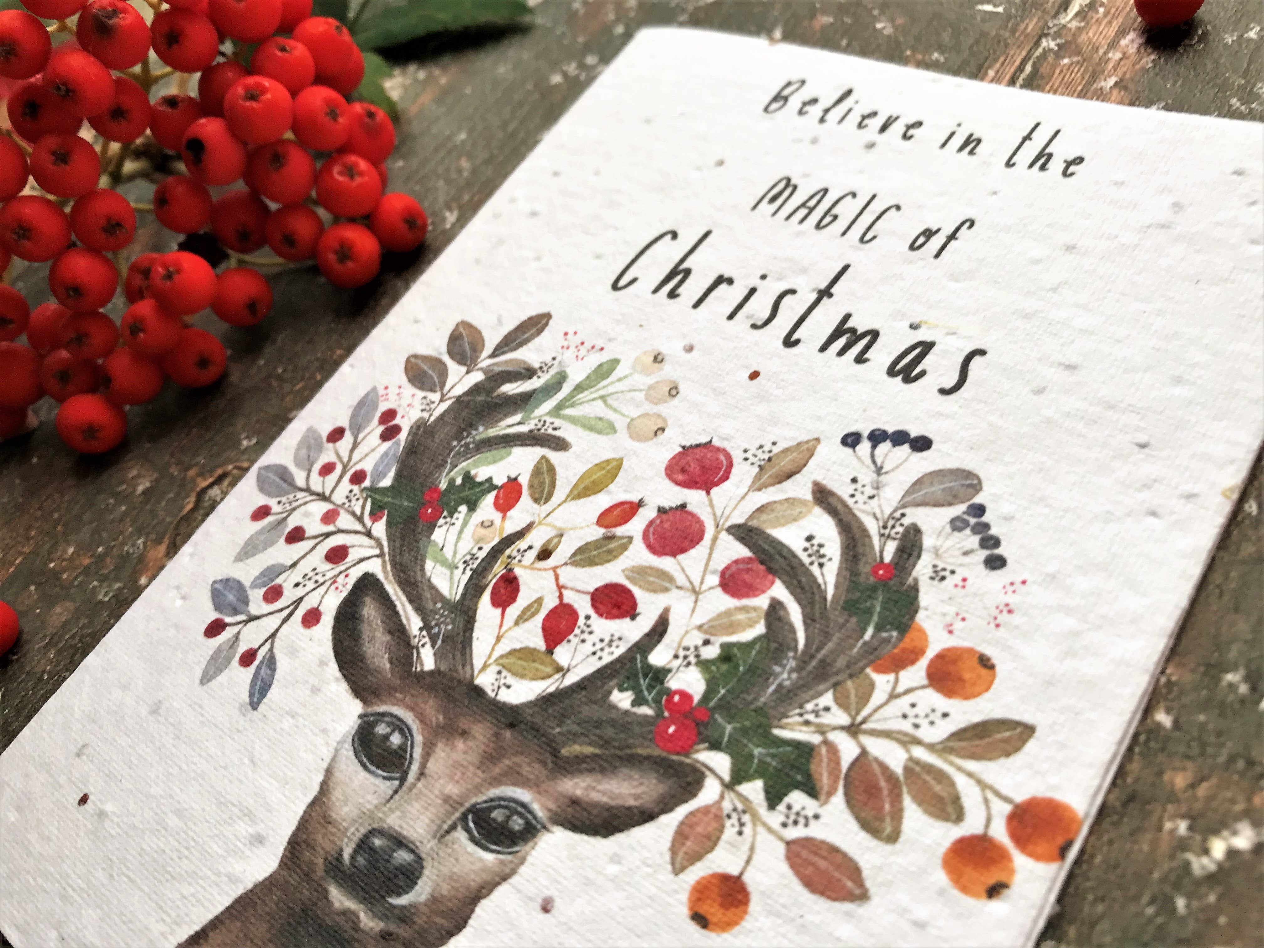 A textured white Christmas greeting card featuring a watercolor deer with antlers decorated with red berries and green leaves, with the text 'Believe in the magic of Christmas' written on it. The card is styled on a rustic wooden surface next to a cluster of red berries.