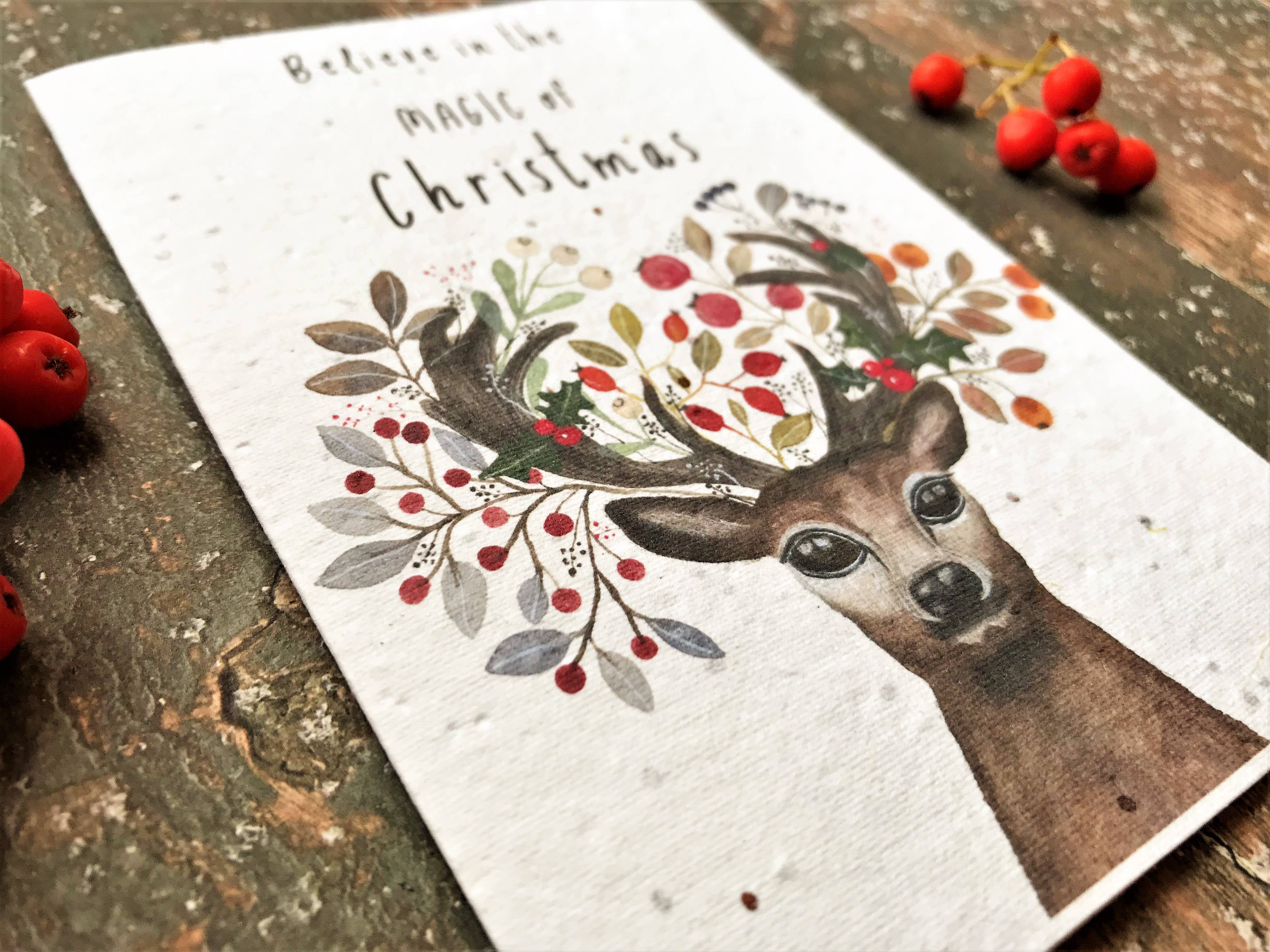 A white Christmas card with a watercolour deer illustration and the phrase 'Believe in the Magic of Christmas,' styled on a rustic wooden surface with red berries around it.