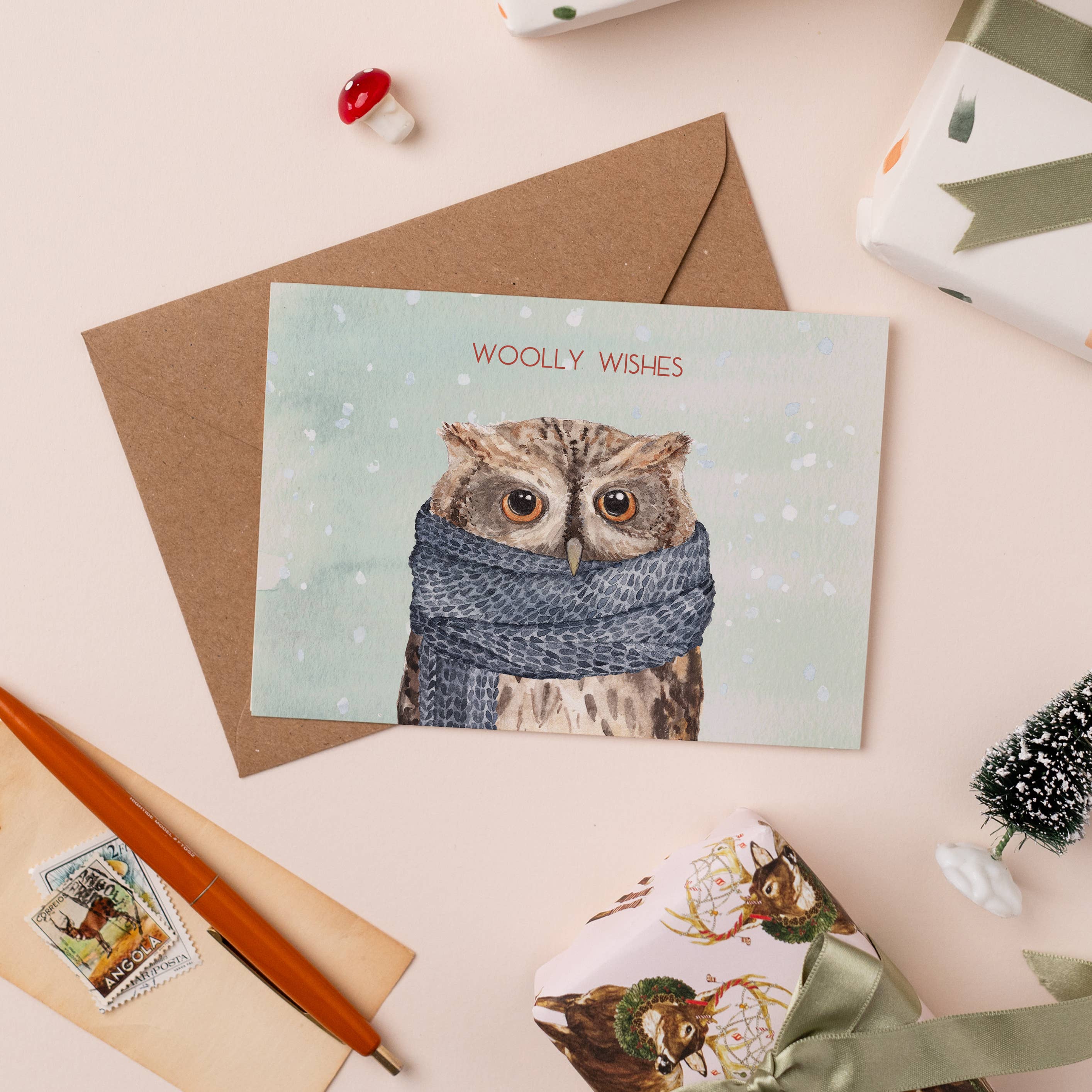 Greeting card with owl wearing a scarf and 'Woolly Wishes' text, surrounded by stationery items on a light background.