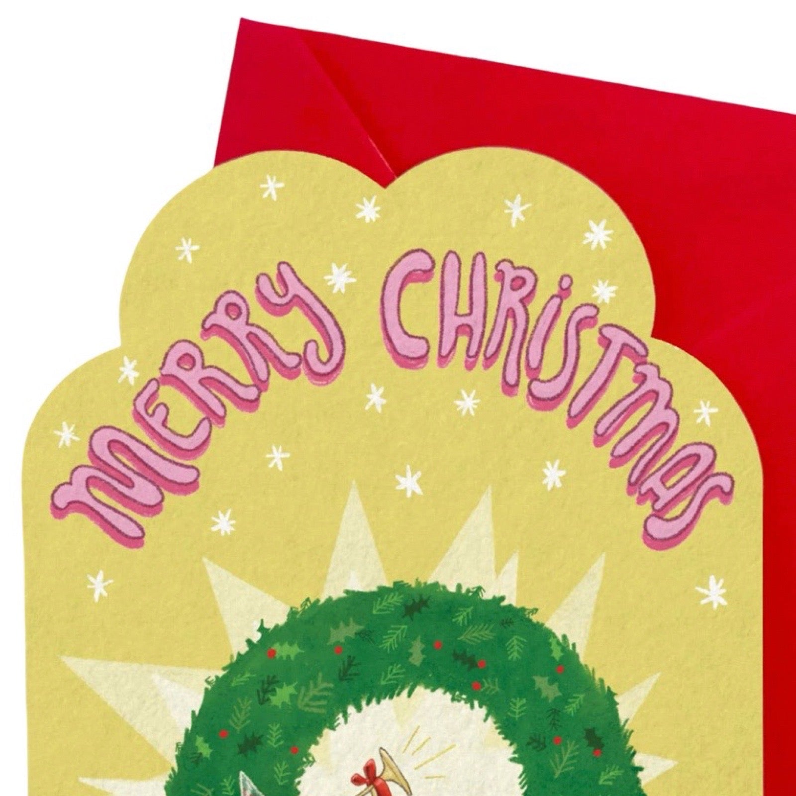 A festive Christmas greeting card with a scalloped yellow front featuring the words “Merry Christmas” in pink script, a green wreath at the bottom, and a red envelope partially visible behind it on a white background.