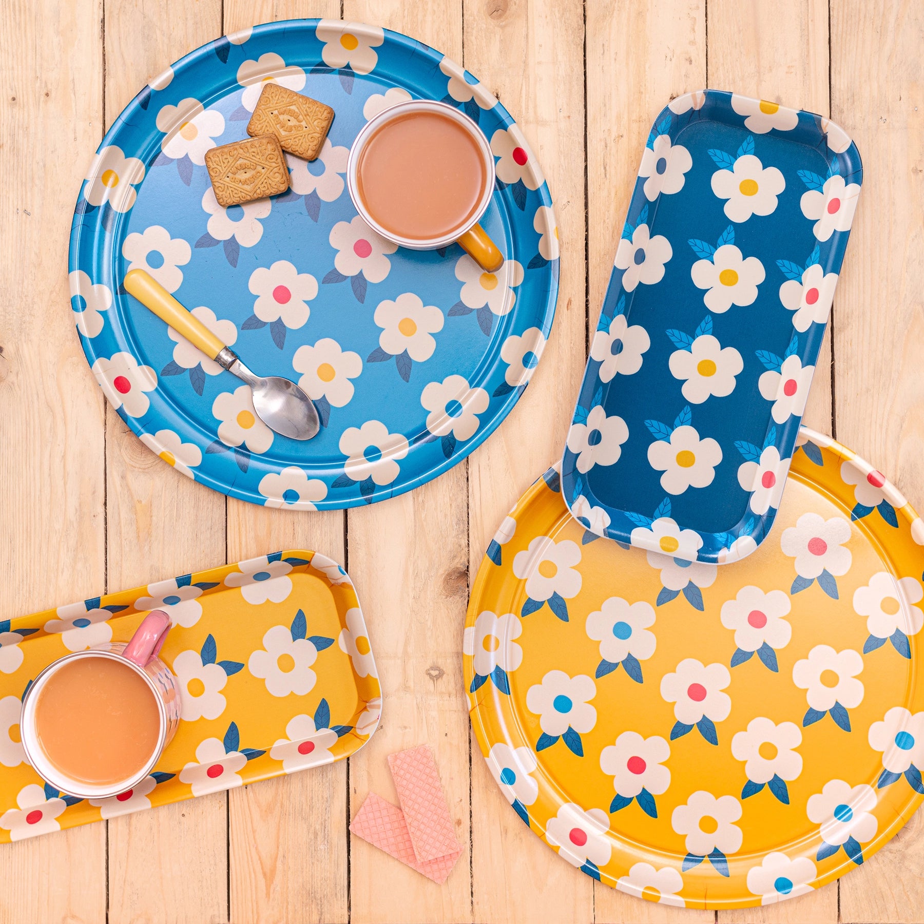 A blue circle floral tray, yellow circle floral tray, rectangle floral blue and yellow trays, on a wooden background with cups of tea, biscuits and spoons.