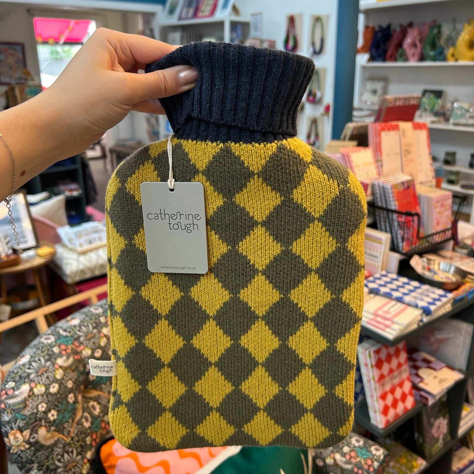 A hand holds a hot water bottle covered in a knitted cover with yellow and dark olive diamonds and a navy ribbed neck. A tag reading 'catherine tough' is visible, with a shop interior in the background.