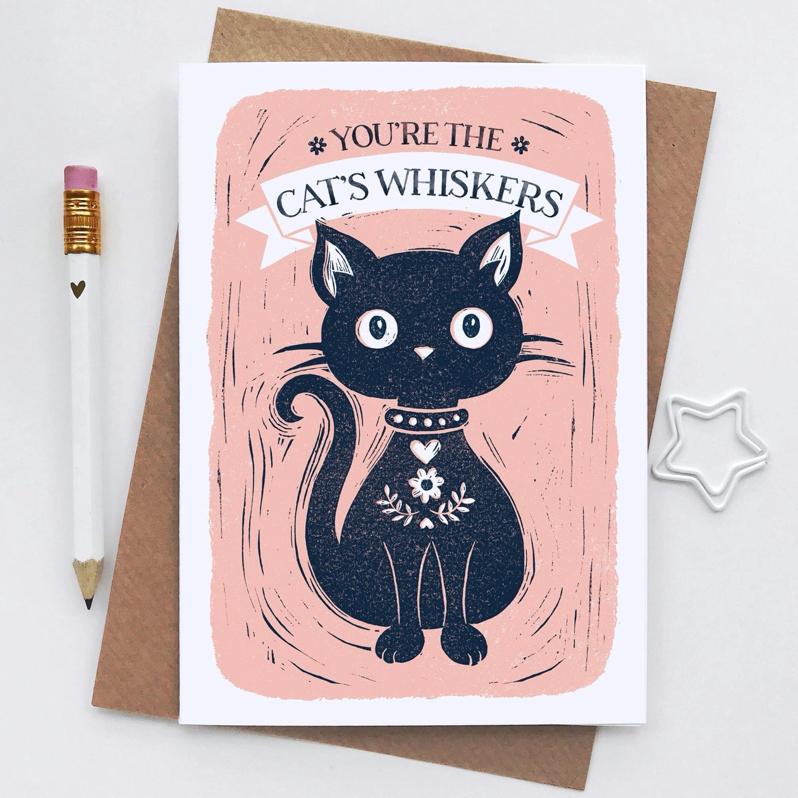 Greeting card with a black cat illustration and text on a pink background, next to a a pencil and star-shaped object.