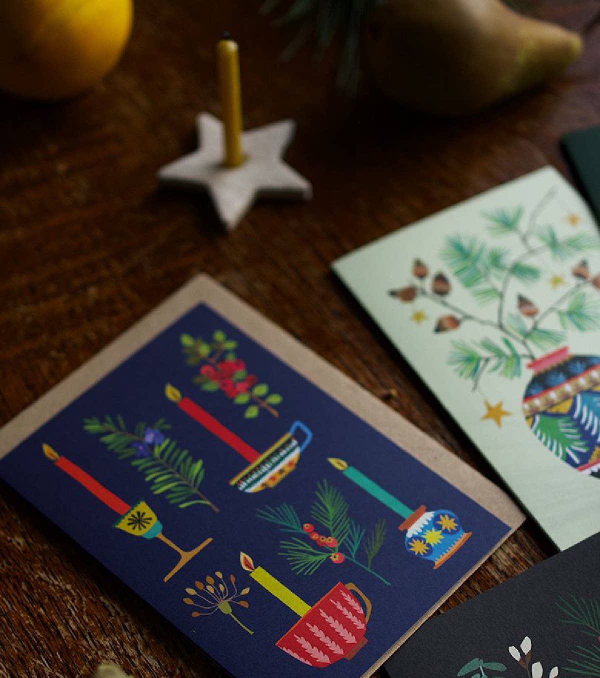 A set of illustrated holiday greeting cards arranged on a rustic wooden table. The most prominent card in the foreground has a dark navy blue background with colorful drawings of candles, evergreen branches, pinecones, and a mug.