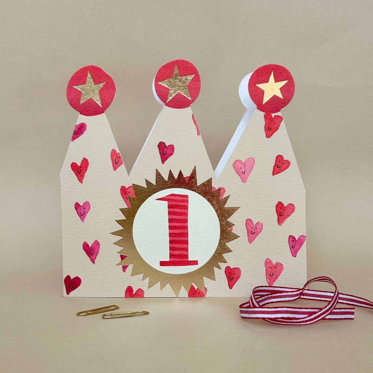Decorative birthday crown with number 1, hearts, and stars on a beige background.