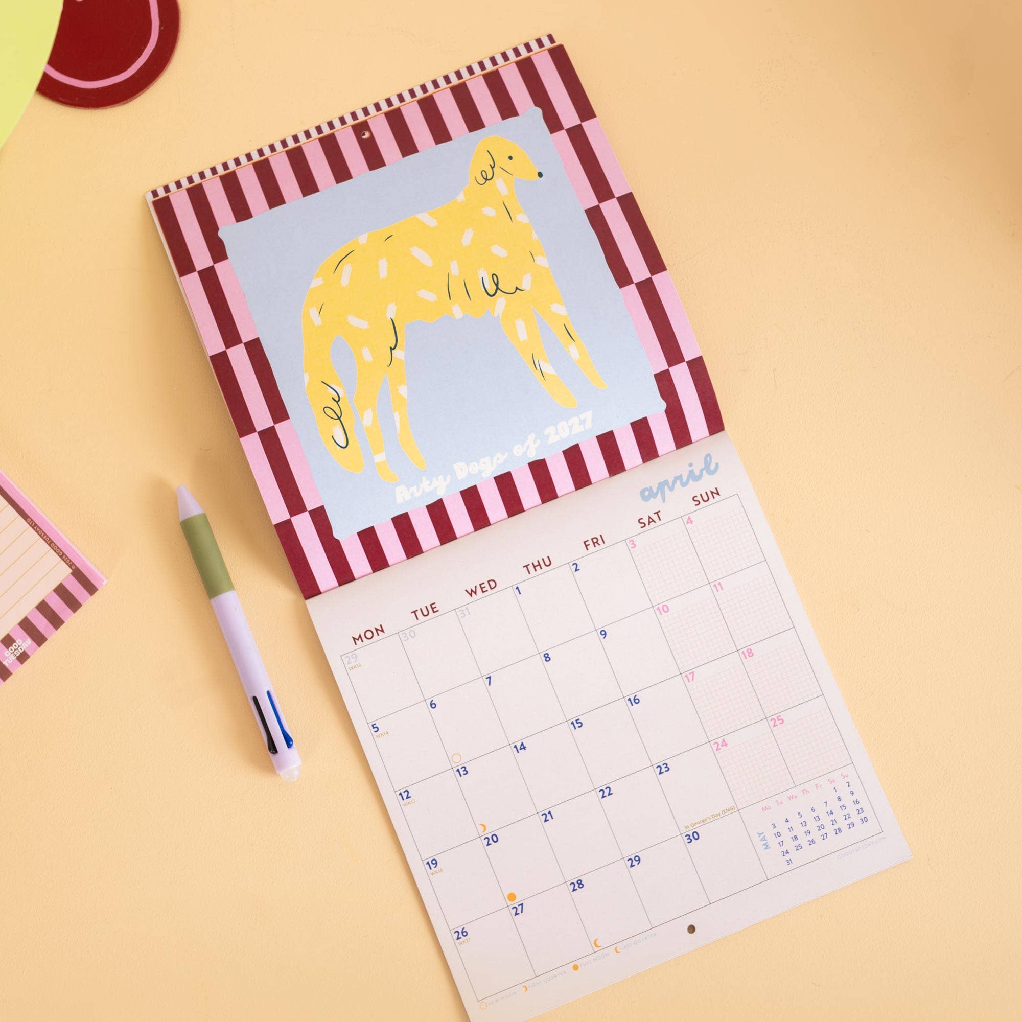 A spiral-bound desk calendar featuring a yellow dog illustration on the cover, styled on a pastel yellow surface with a pen and a small notepad nearby.