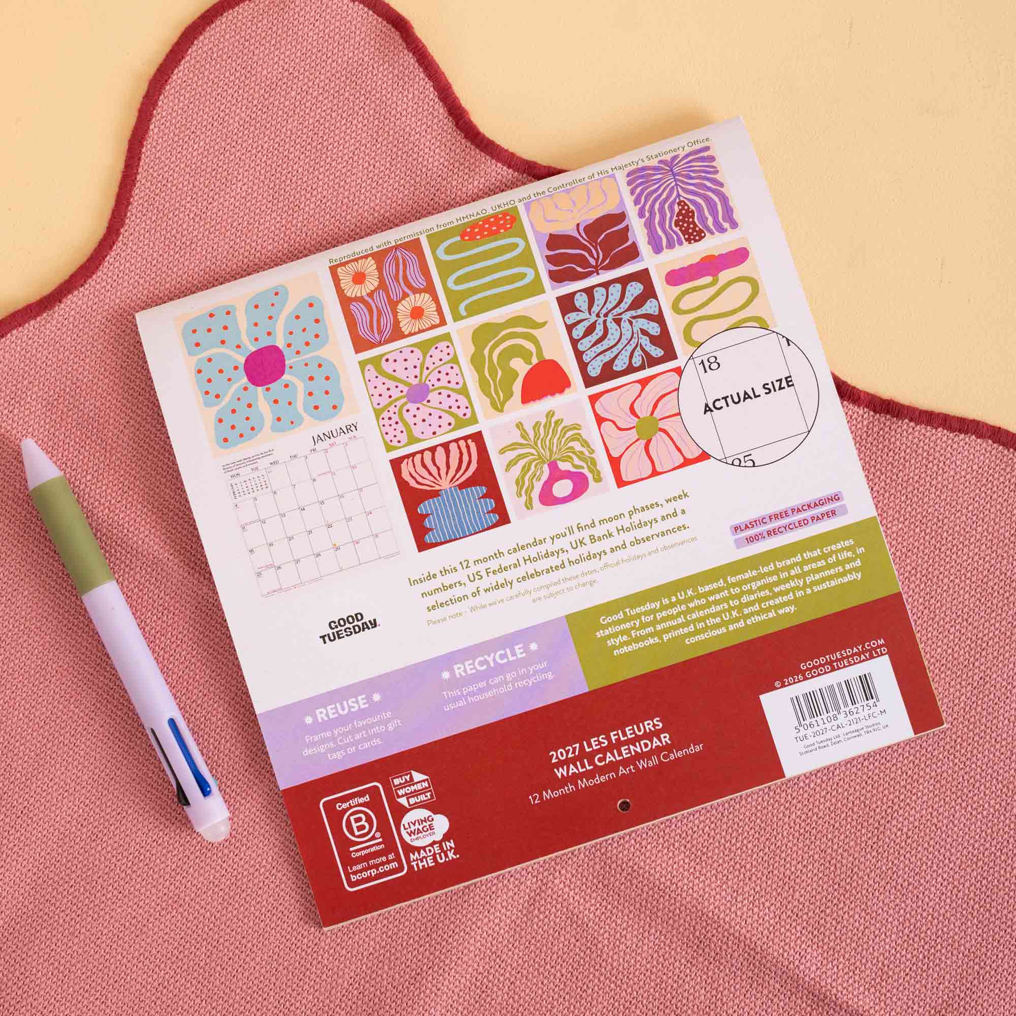 A 2027 Les Fleurs Mini Calendar is shown from its back side, featuring a colourful grid of flower illustrations and product details. It is placed on a textured pink fabric next to a pastel green and white pen.