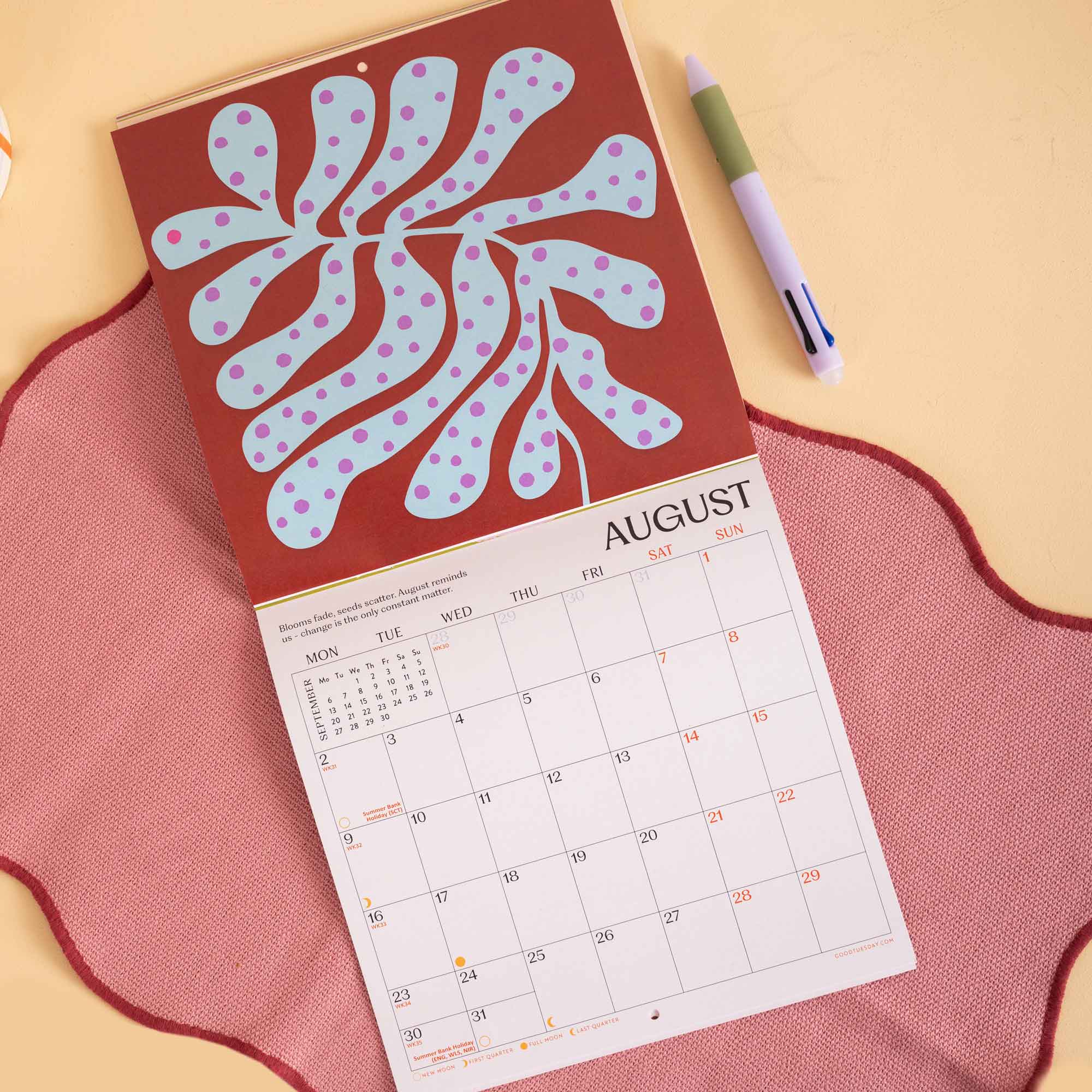 A wall calendar page for August is laid on a pink fabric surface, showing a large abstract leaf-like illustration in pale blue and lavender on a reddish-brown header. A green-and-white pen rests beside it on a warm beige background.