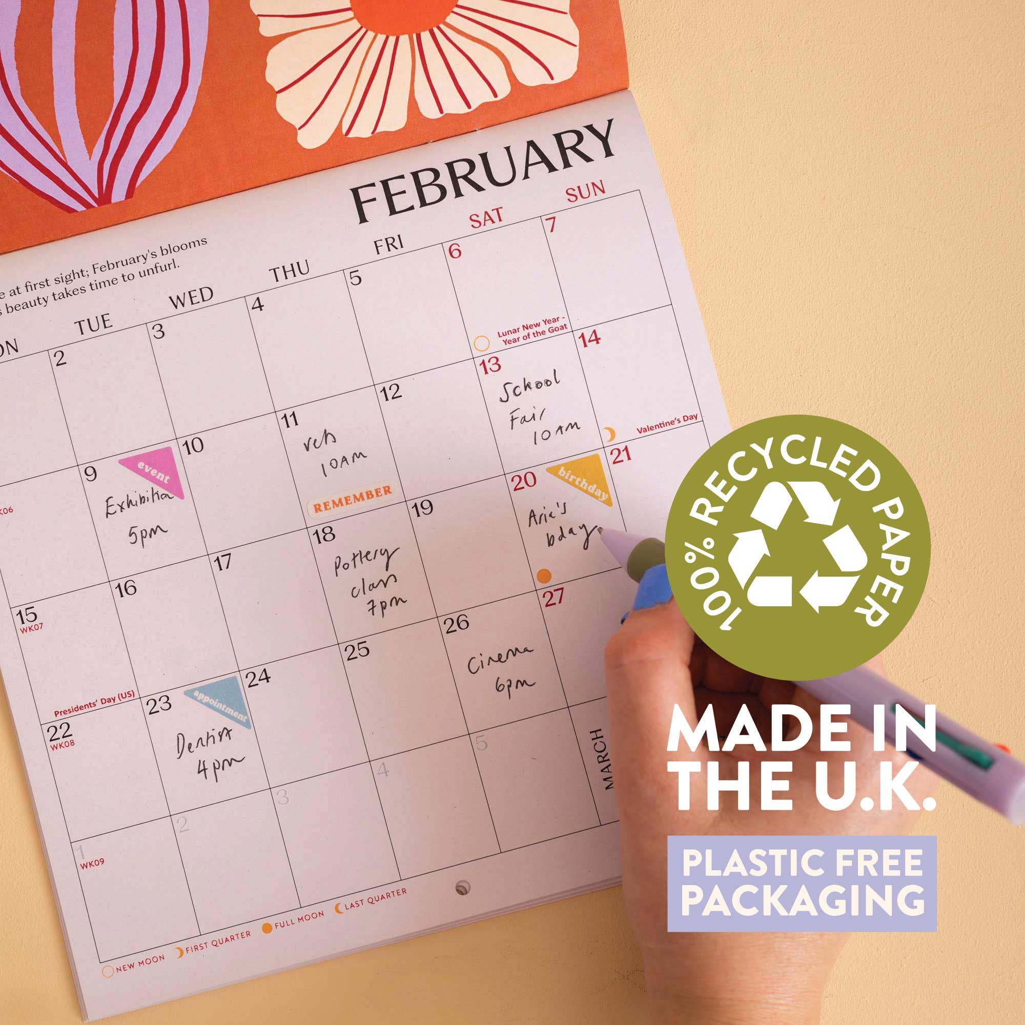 A close-up of a February planner page from The Happy Planner, held by a hand with a green pen. The page features a colorful floral header and a grid for daily planning.