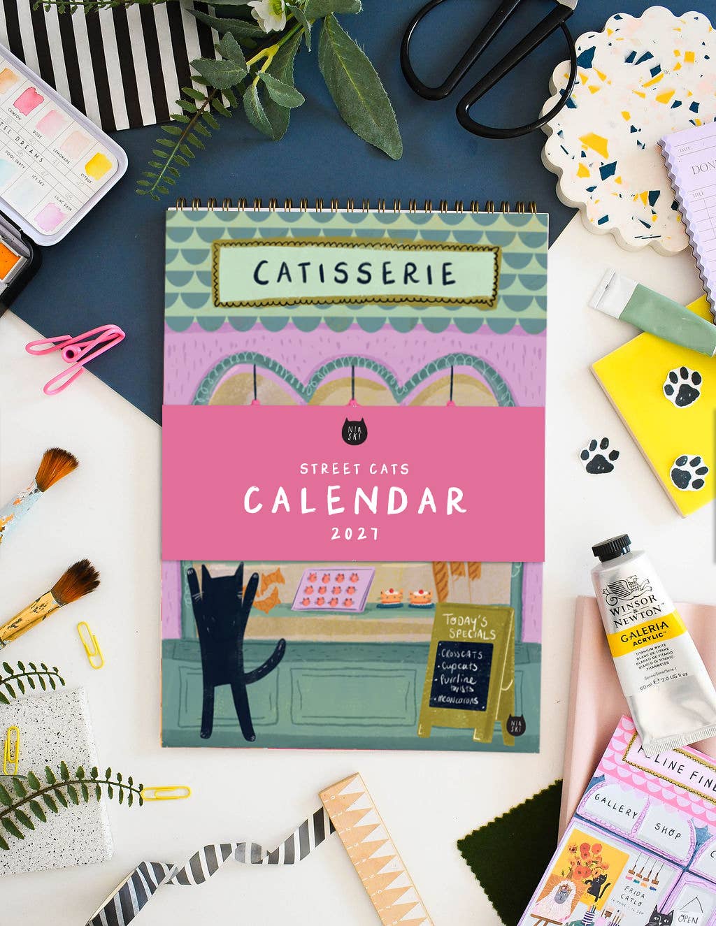 A spiral-bound 'Catisserie Street Cats Calendar 2027' on a desk surface surrounded by art supplies, brushes, paper clips, a paint tube, and patterned accessories. The calendar cover features an illustrated storefront scene with a black cat silhouette and pastel colours.