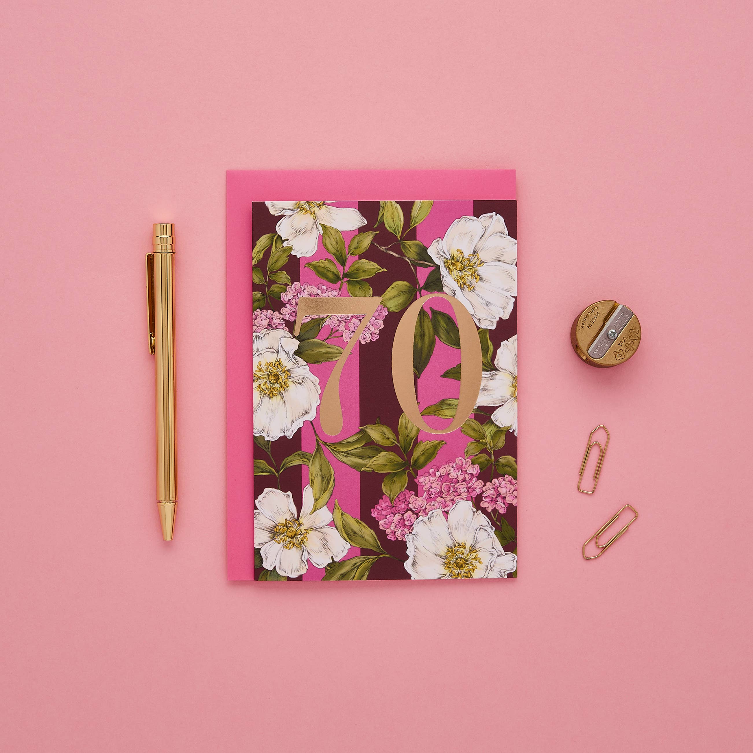 A flat-lay of a floral '70' birthday card with a pink envelope, styled on a pink background with a gold pen, paper clips, and a sharpening stone.