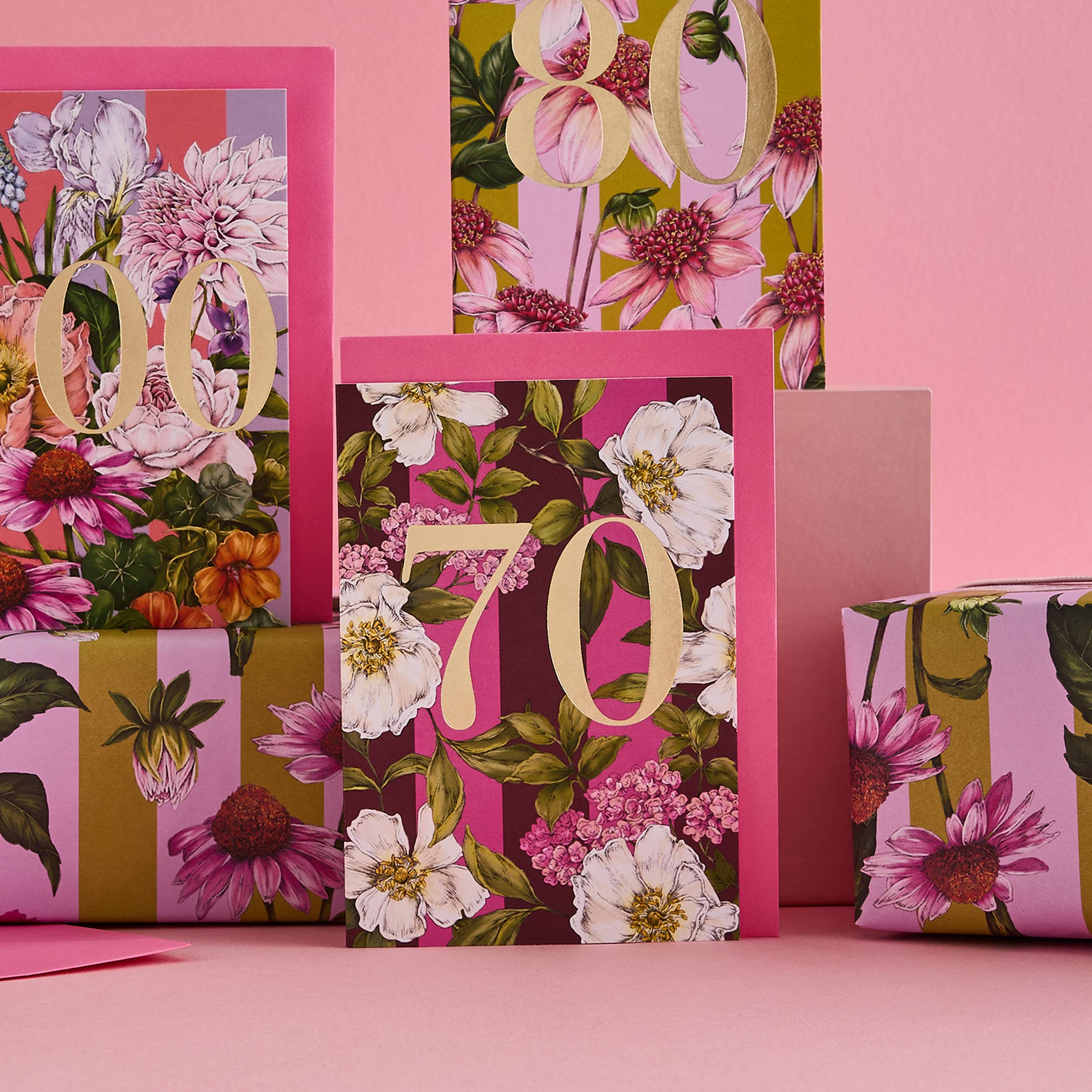 A set of floral birthday cards and matching wrapped gift boxes in a coordinated pink, gold, and botanical print design, arranged on a pink surface against a pink background.