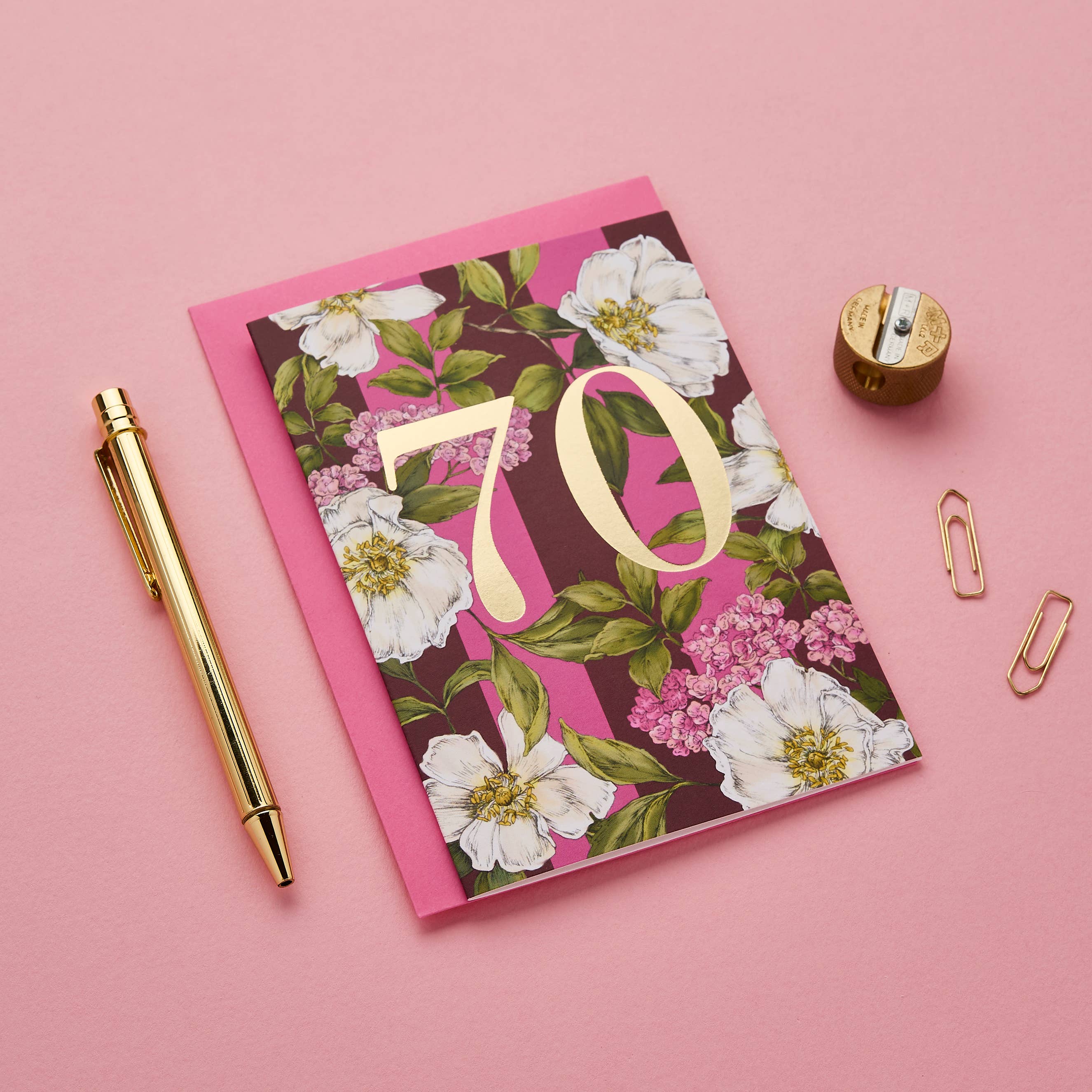 A floral 70th birthday card with a pink envelope, styled on a pink background with a gold pen, gold paper clips, and a gold pencil sharpener.