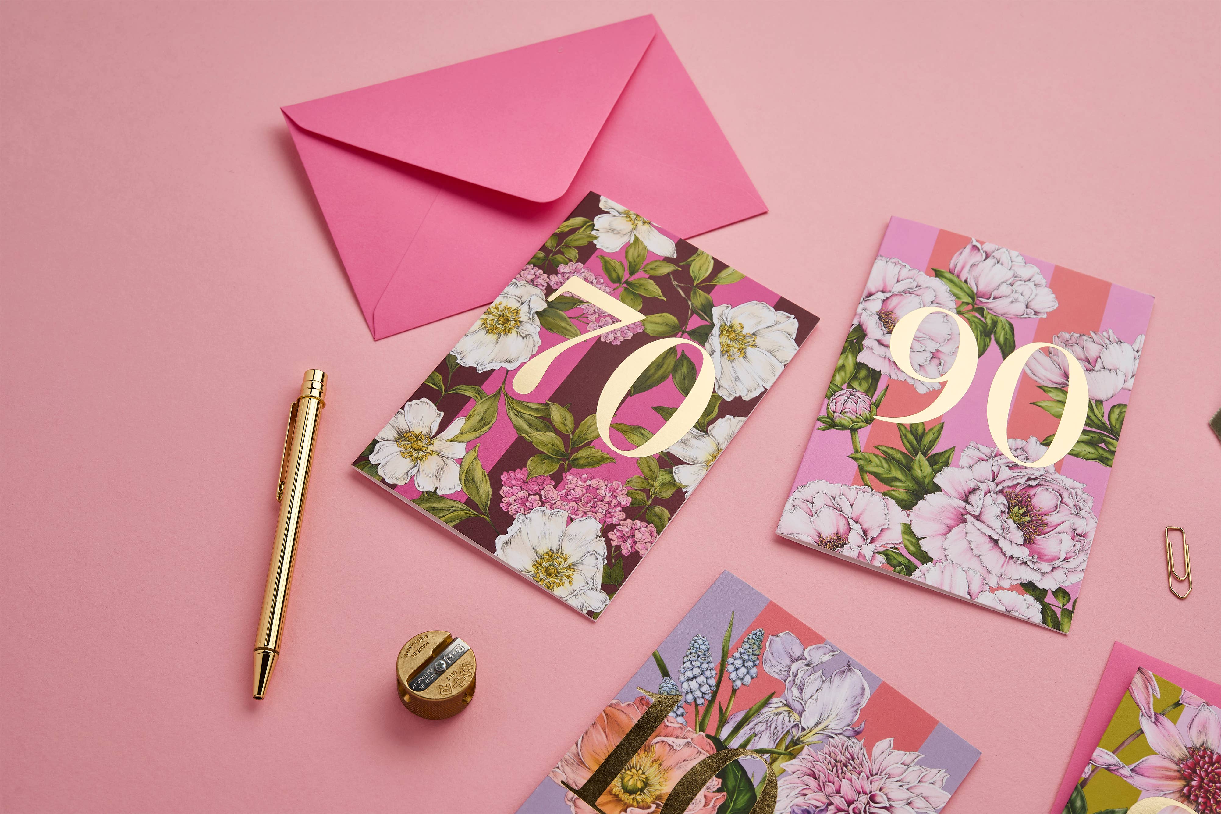 A styled flat-lay of floral greeting cards with gold numbers, a pink envelope, a gold pen, a small gold object, and paperclips on a pink background.