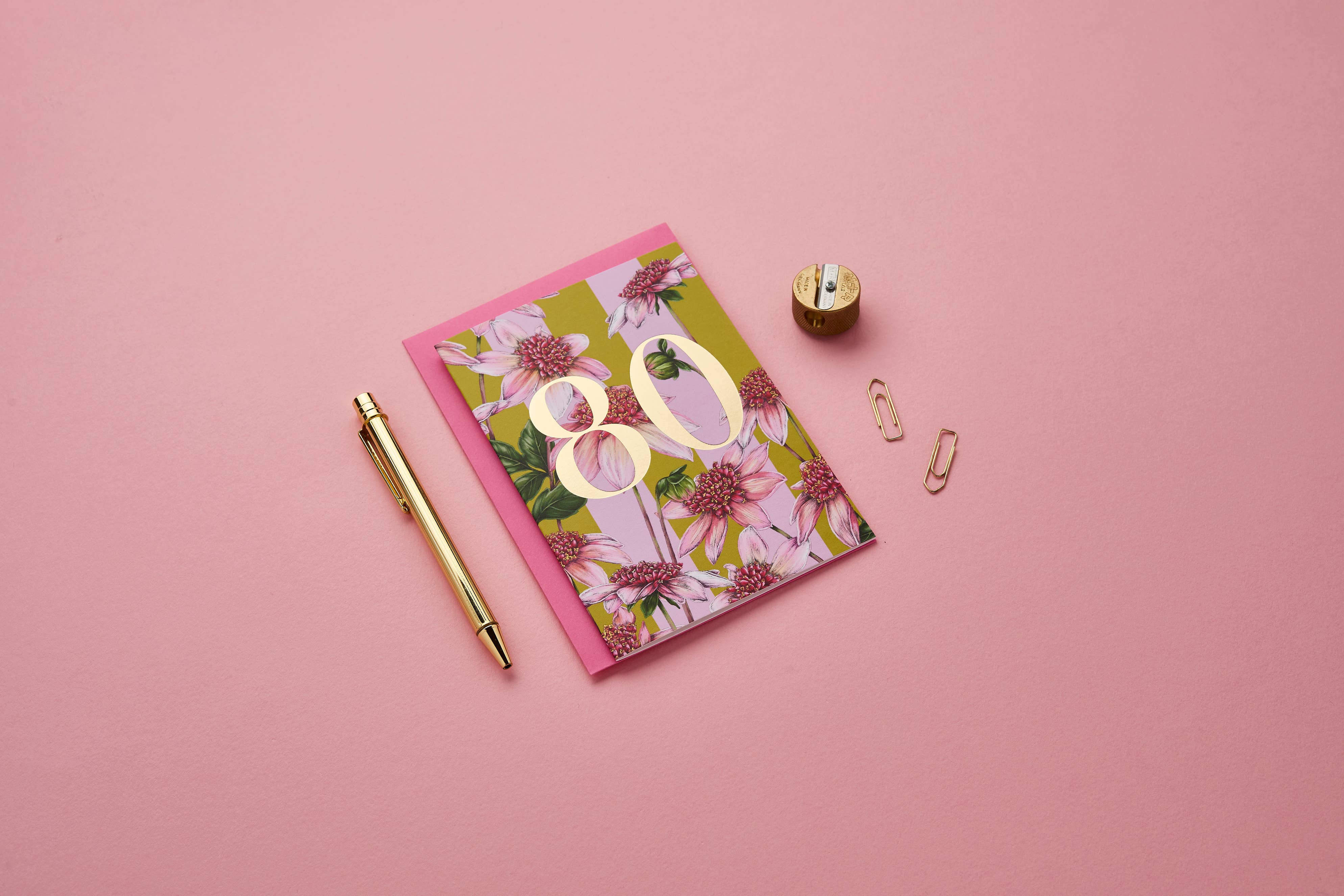 A flat-lay of a floral '80th birthday' greeting card with gold foil numbers, styled with a gold pen, a gold pencil sharpener, and paper clips on a solid pink background.