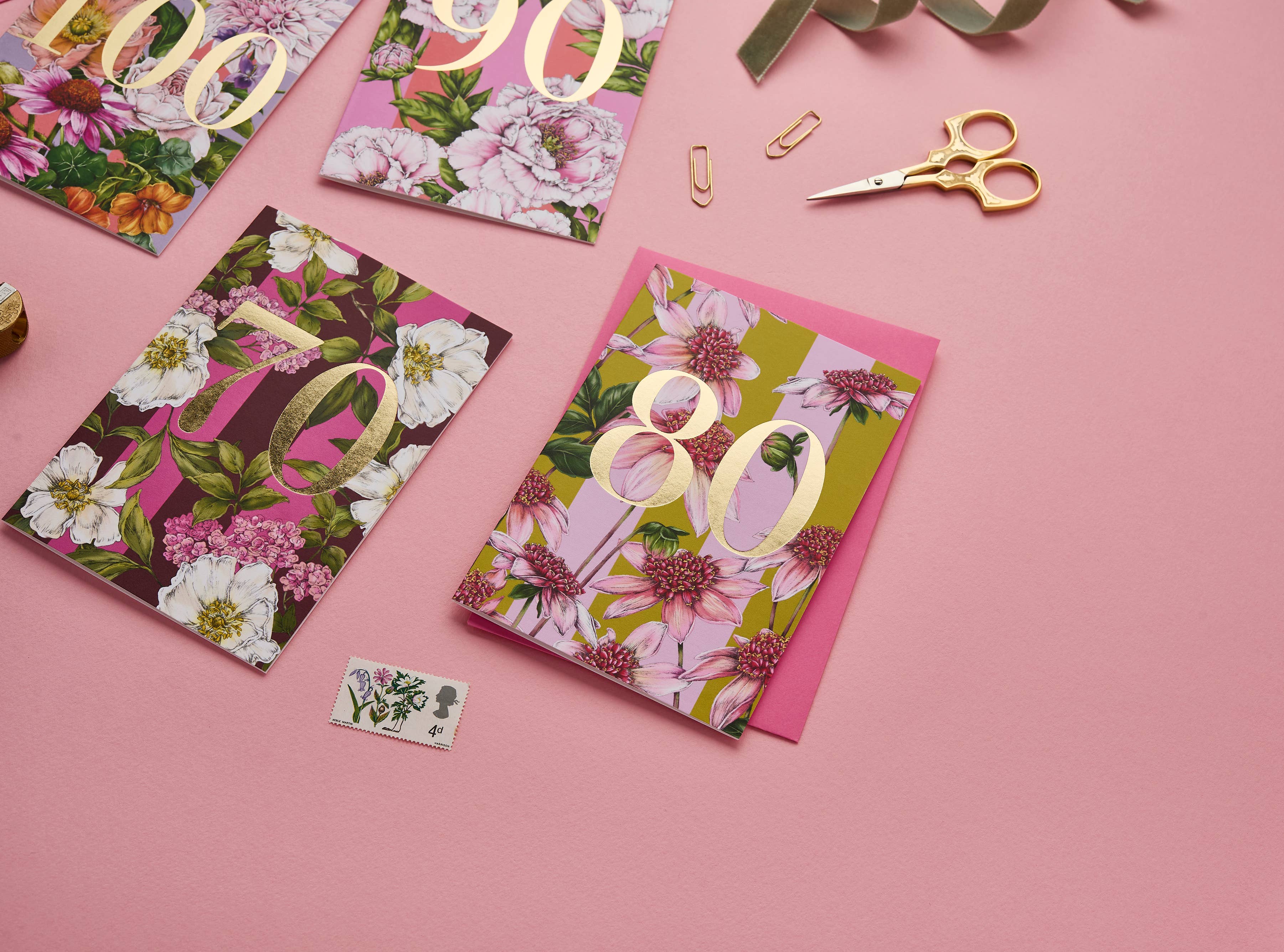 A styled flat-lay of floral milestone greeting cards on a pink background, with gold foil numbers 80, 90, and 100, plus gold scissors, paper clips, ribbon, and a small botanical stamp.