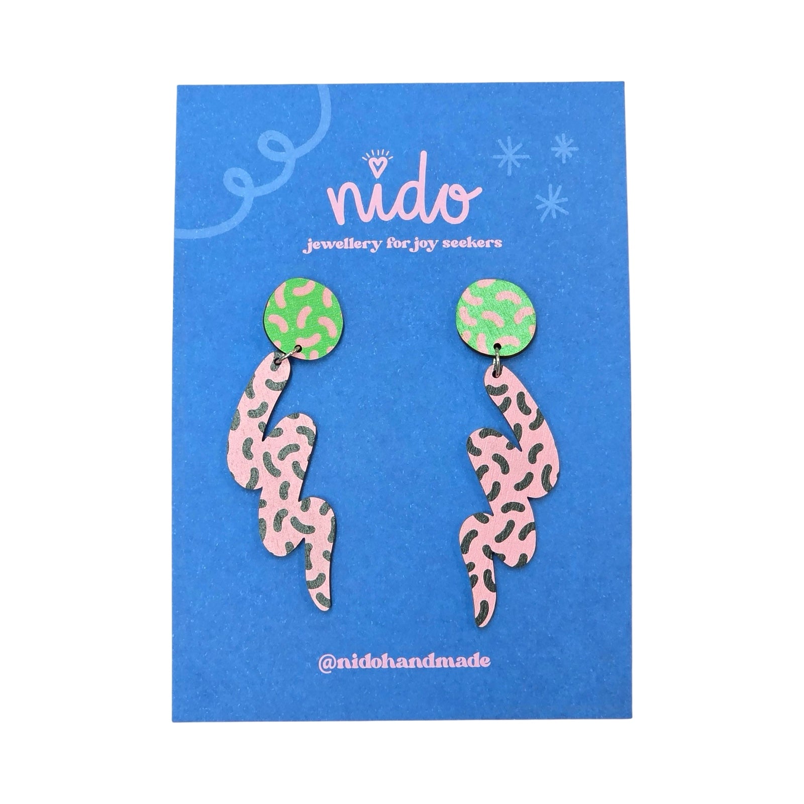 Wooden earrings featuring green circular studs with pink dashes with a large pink and black dashed squiggle hanging underneath, presented on a branded blue backing card.