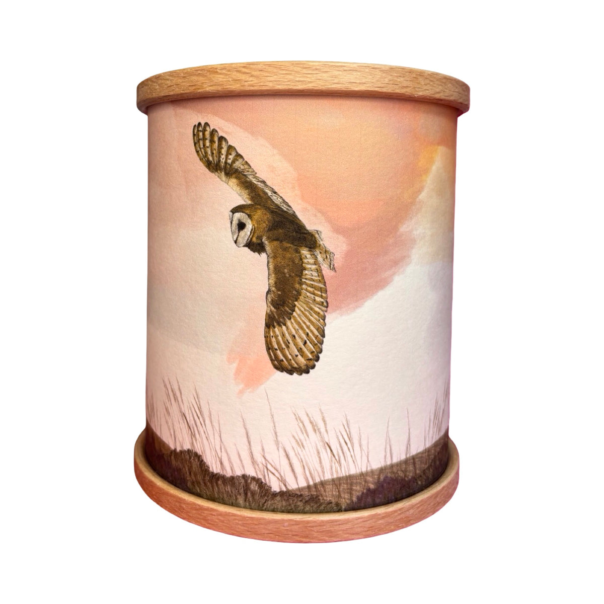 A cylindrical wooden candle holder featuring a printed illustration of a barn owl in flight against a soft peach and pink sky with grass at the bottom. The wood top and base are light natural oak.