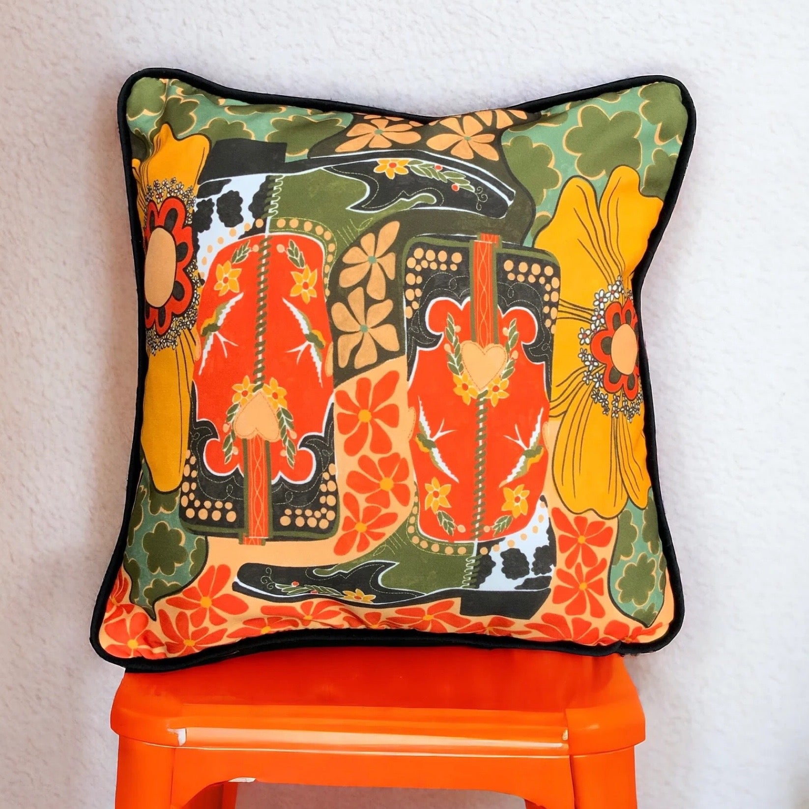 A cushion cover featuring a pair of cowboy boots surrounded by flowers, all in retro tones, sat on an orange chair with a white wall behind.