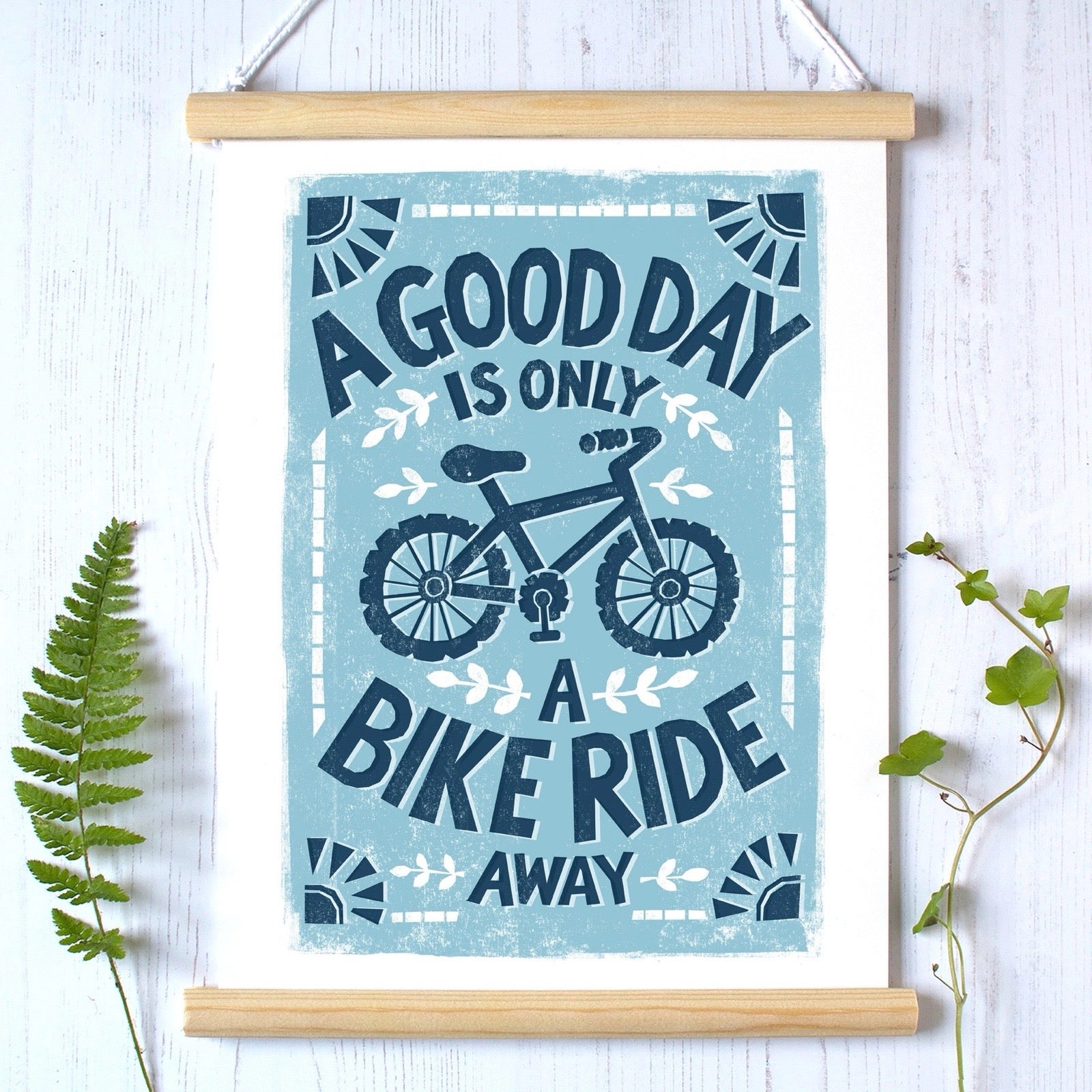 Poster with bicycle design and text hung up by a wooden frame against a white background with leaves.