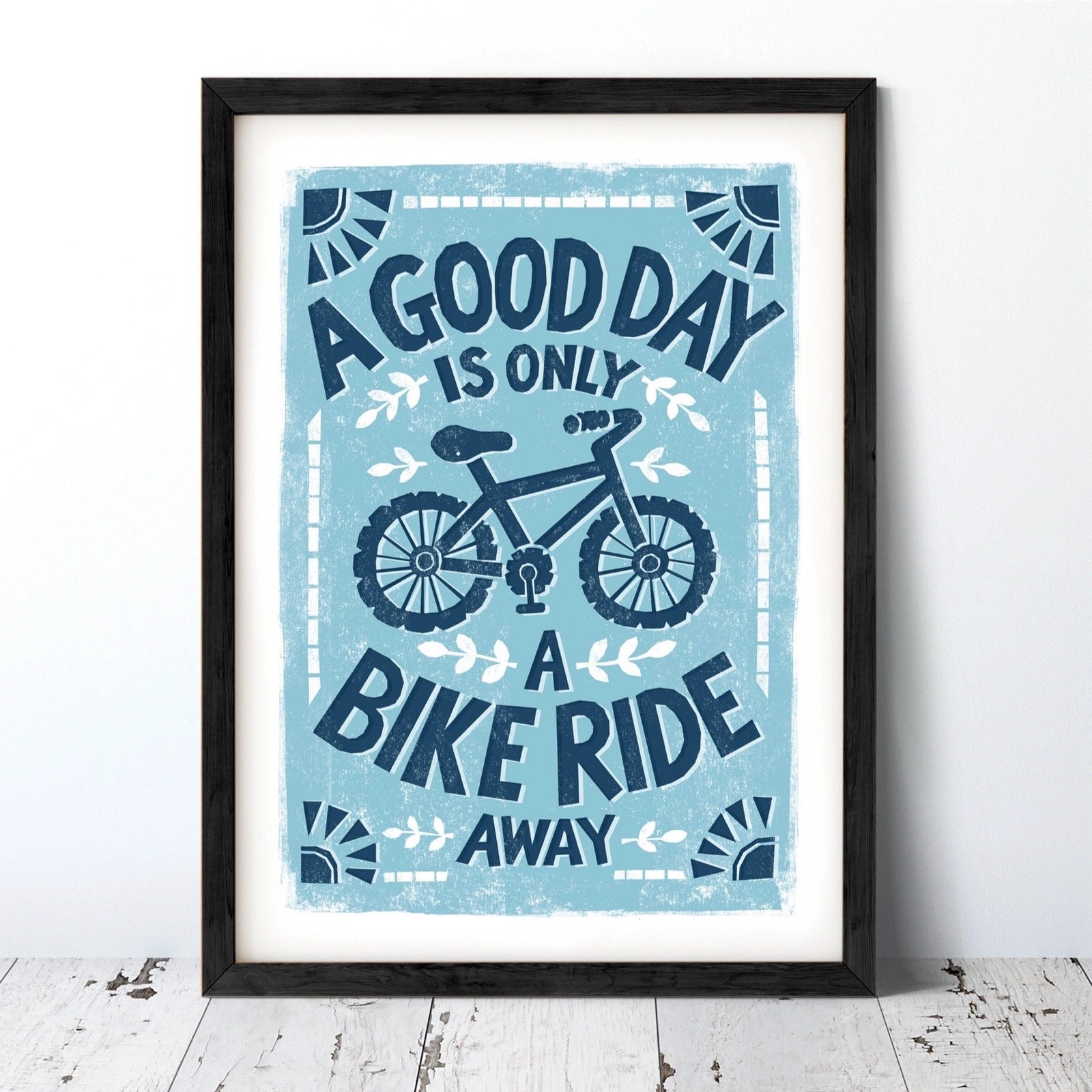 Framed poster with a bicycle design and text on a light blue background, against a wall.