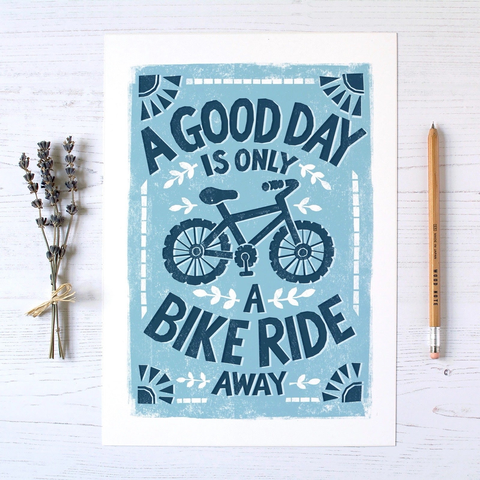 Poster with bicycle design and text 'A good day is only a bike ride away' on a light blue background, placed on a white surface with a pencil and lavender on the side.