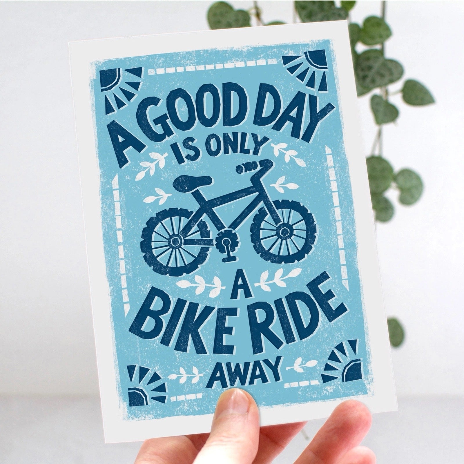 Card with bicycle design and text 'A good day is only a bike ride away' held by a hand, with leaves in the background.