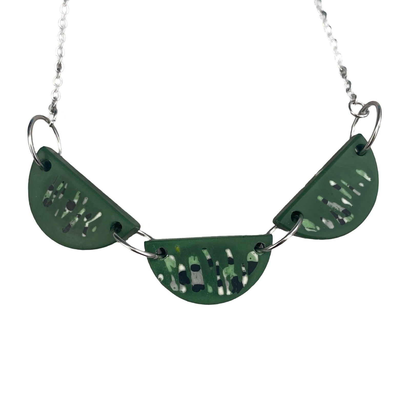 A necklace with three half-moon resin shapes in green with a black and white abstract pattern held together with silver findings on a silver chain.