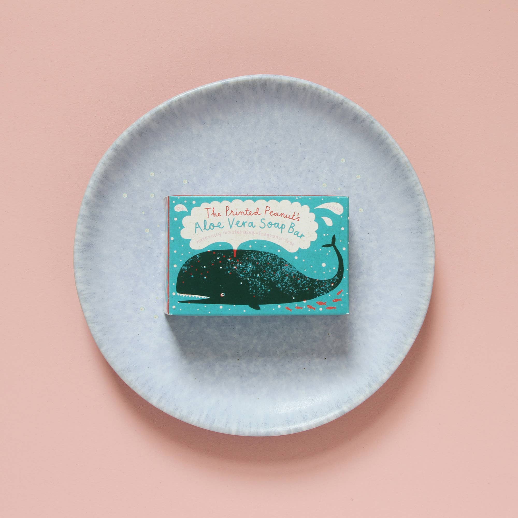 Soap bar with packaging on a blue dish against a pink background.