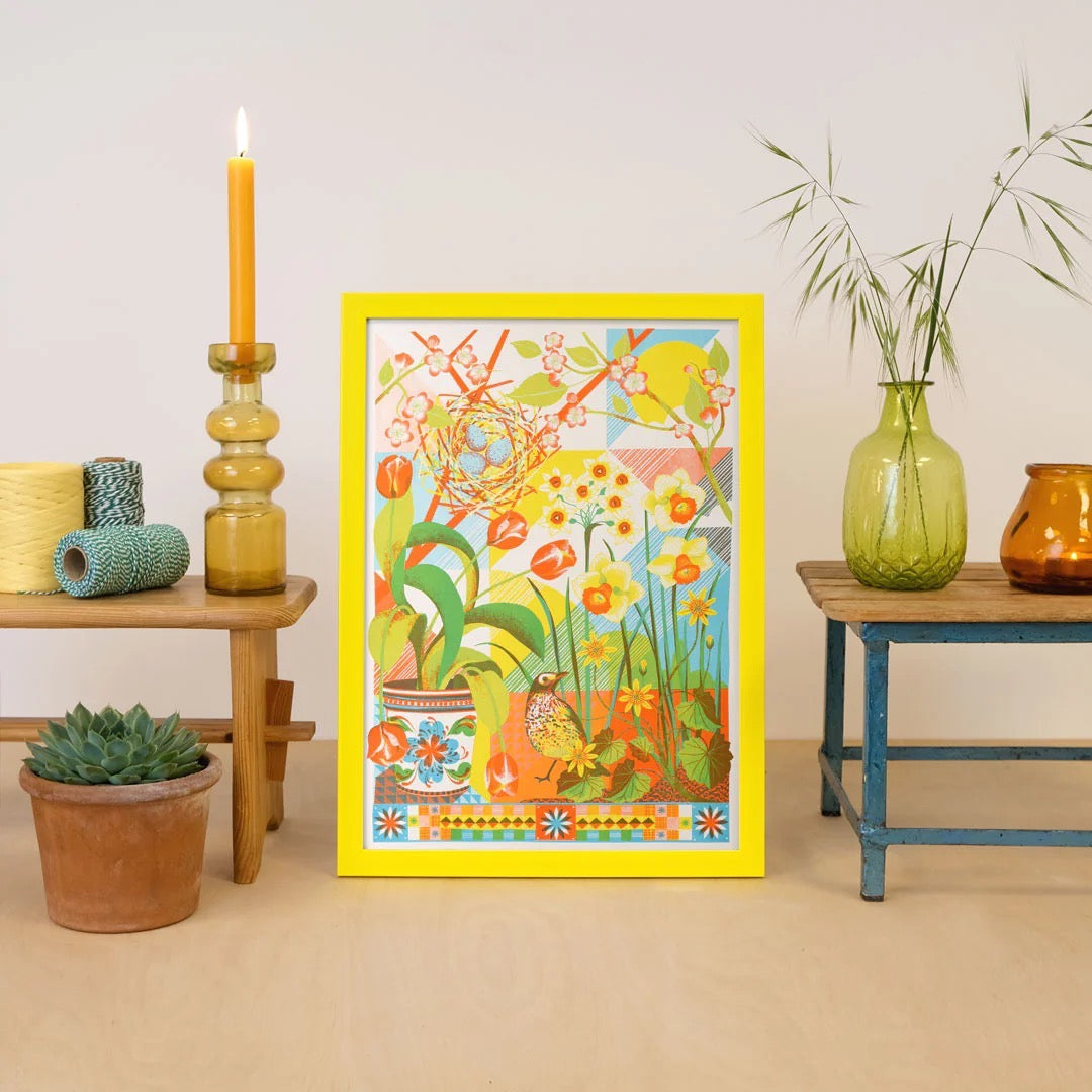 Framed colourful artwork of a garden scene with birds and flowers on a beige wall, next to benches with decorative plants and candles.